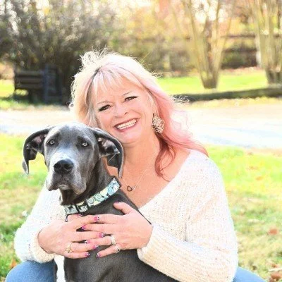 Woman with pink hair smiling, holding a black dog outdoors in a park-like setting.