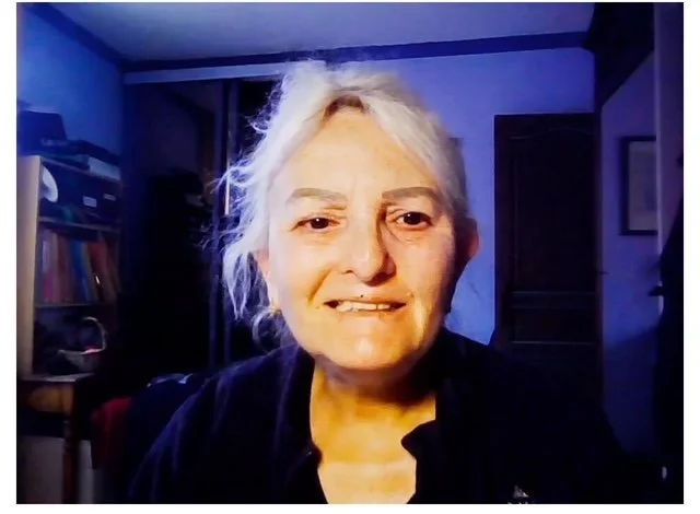 Une femme âgée avec des cheveux gris clair, portant un haut noir, souriante, dans une pièce avec des étagères et des livres.