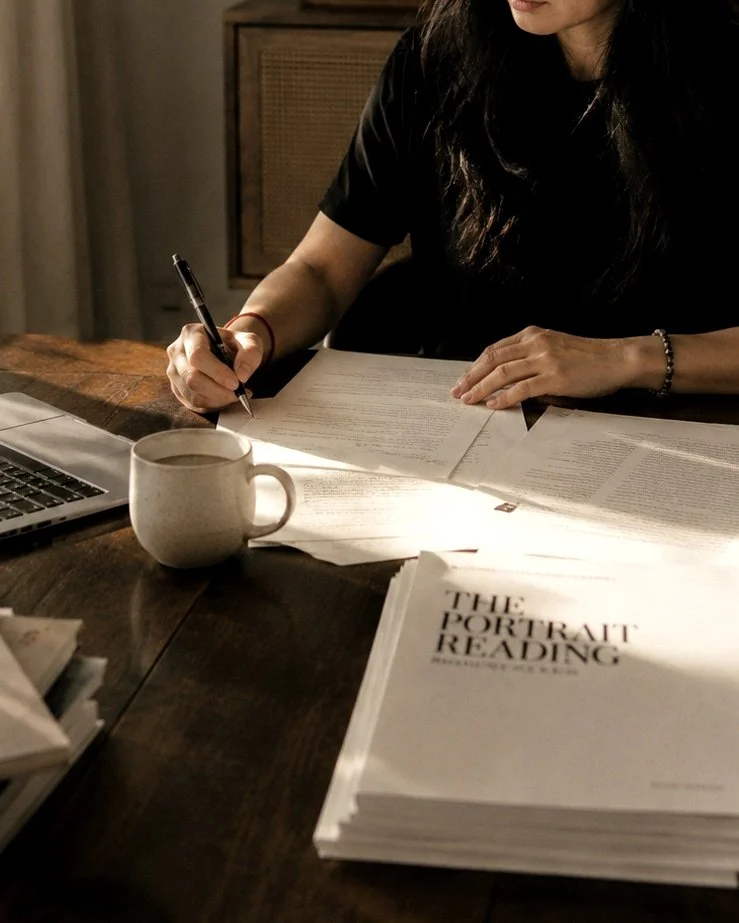 A close-up photograph of a person's hands writing notes on papers at a dark wooden desk, next to a ceramic coffee mug, a laptop, and a printed document titled "The Portrait Reading.