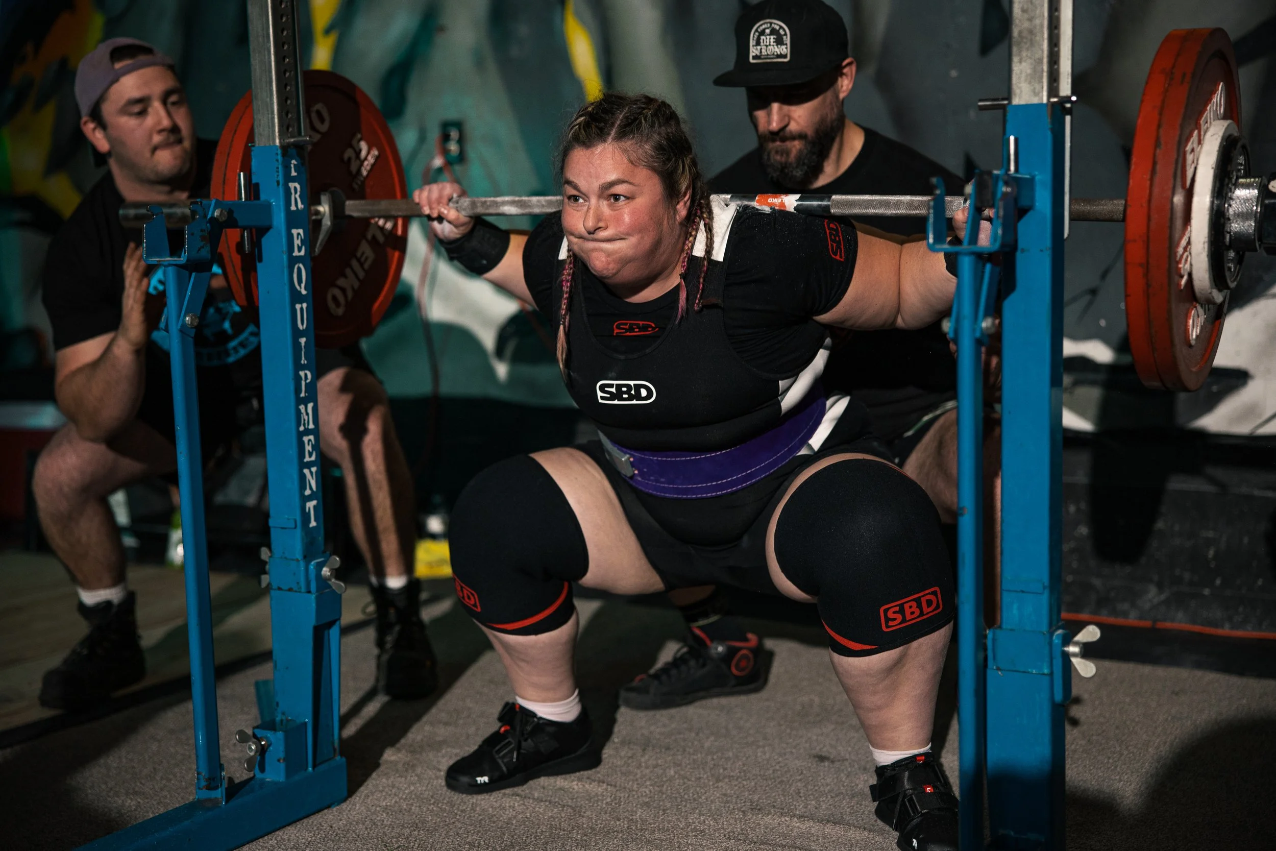 Laura performing a squat exercise with a barbell loaded with orange weight plates, assisted by two spotters at a competition. She is wearing a black athletic outfit with red logos, and her hair is braided.