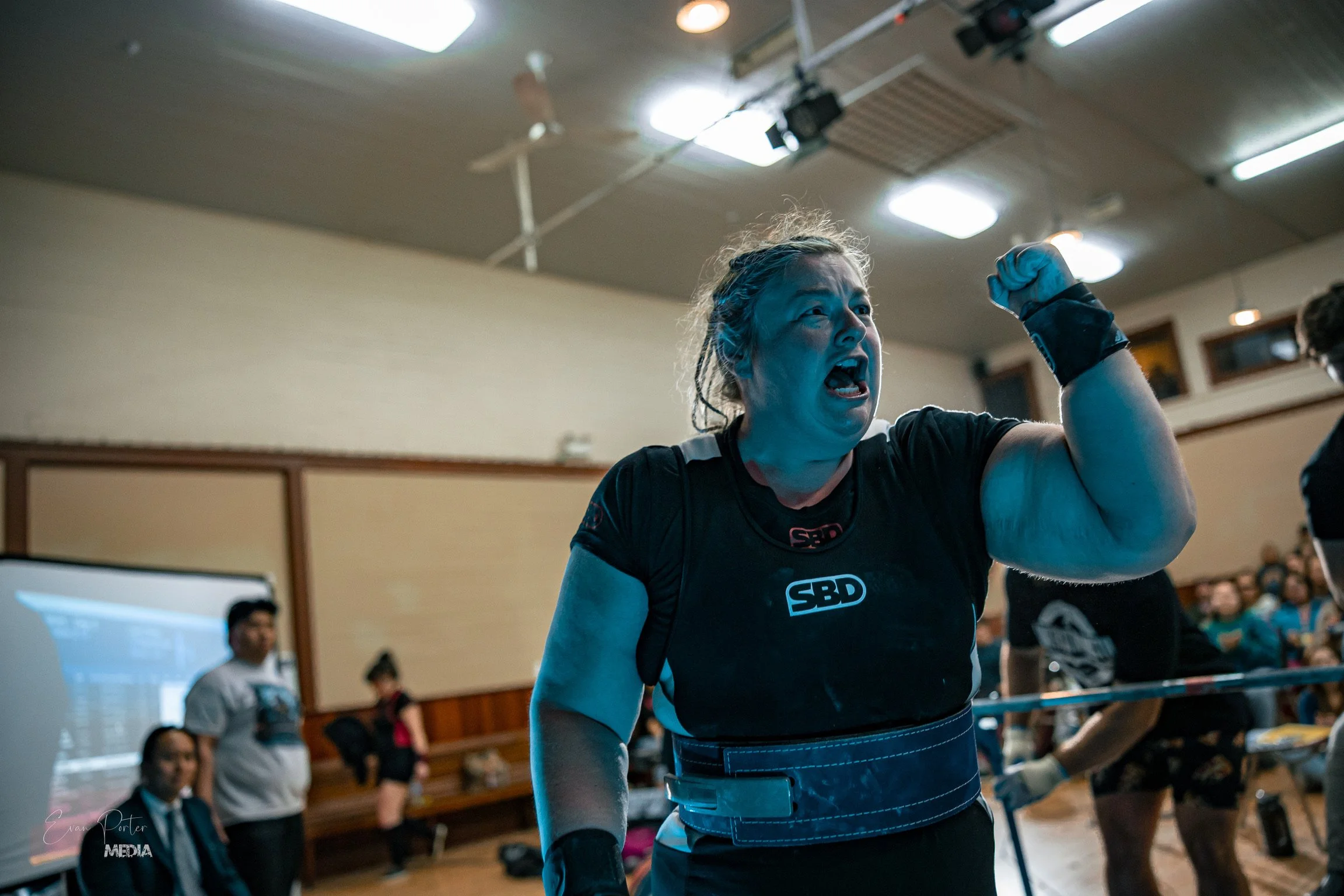 Laura with braided hair and a black shirt cheering in front of an audience in a gymnasium at a powerlifting competition.