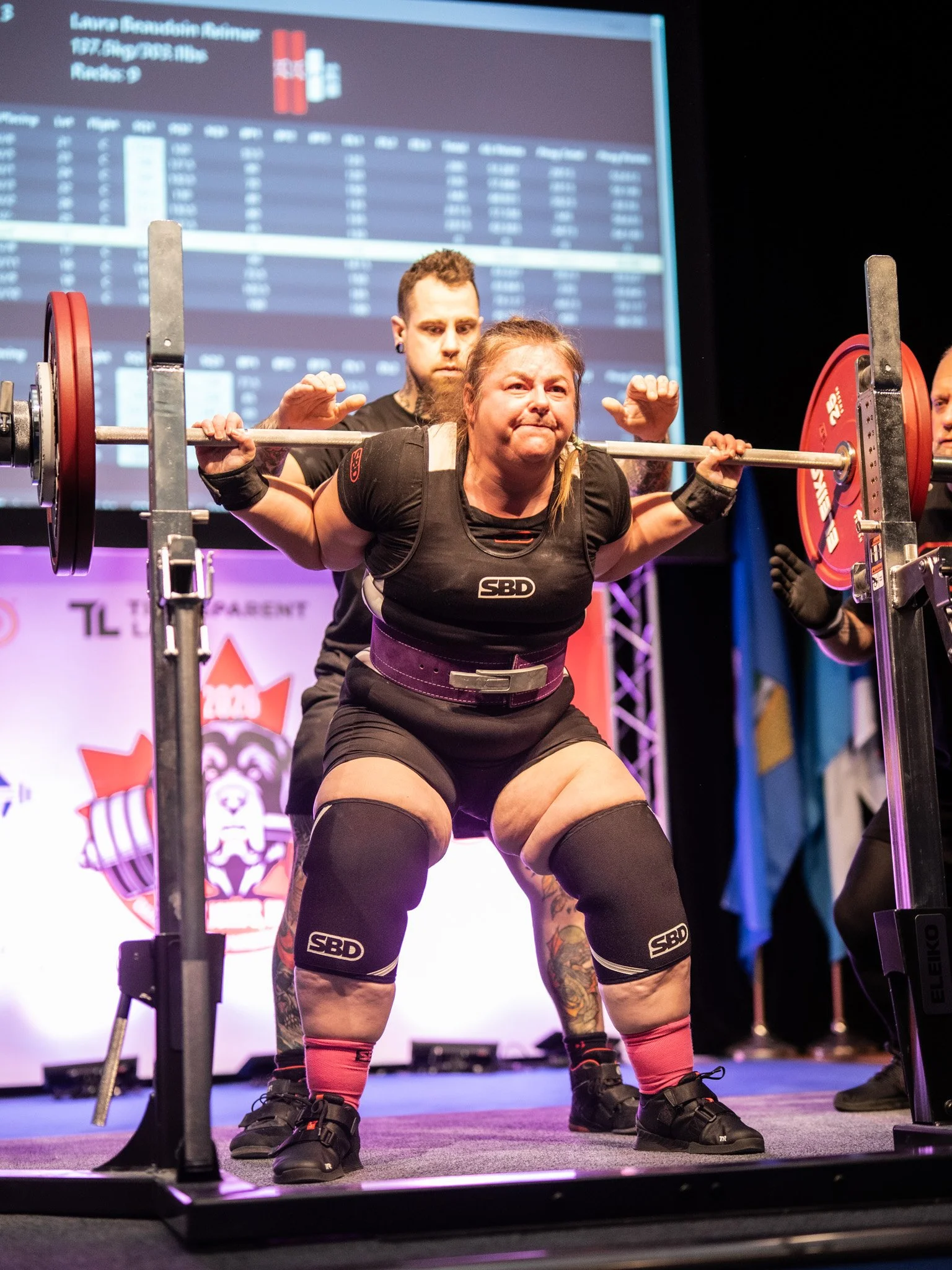 Laura performing a squat with a loaded barbell during a weightlifting competition, with a spotter behind her and a scoreboard in the background.
