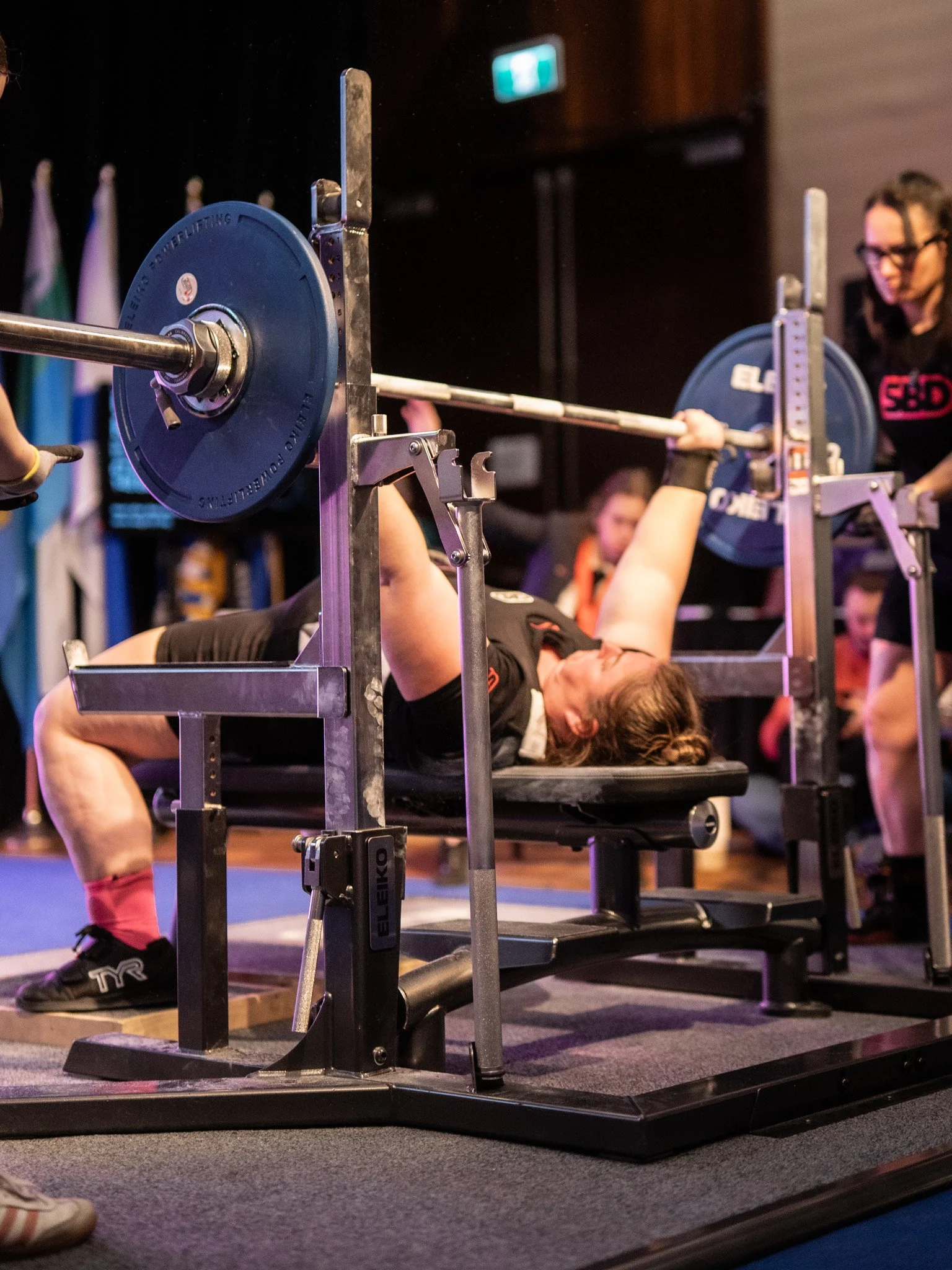 Laura lying on a bench press with a barbell, spotter standing nearby, gym setting with flags in the background.