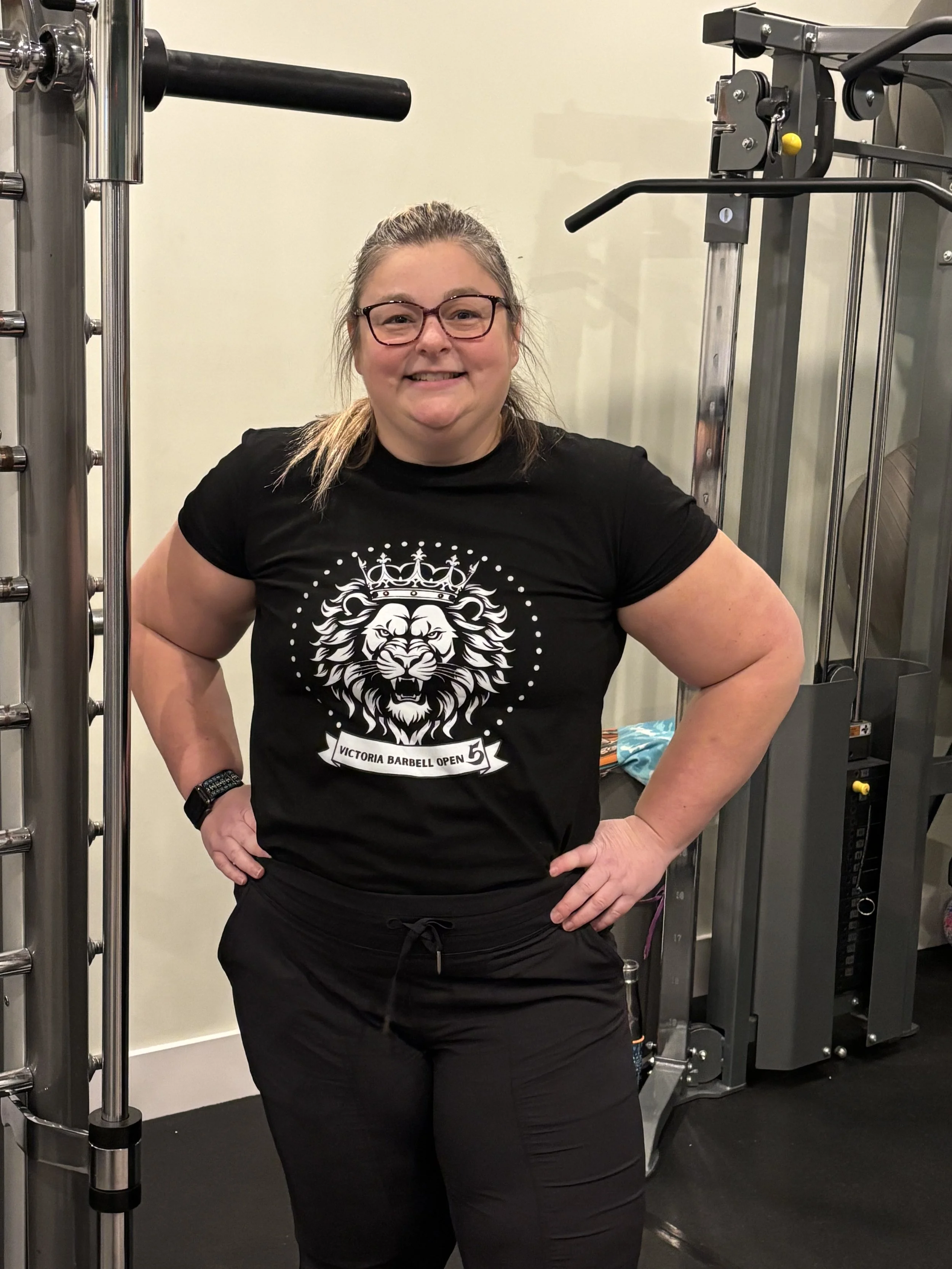 Laura with glasses and a black T-shirt smiling, standing in a gym near workout equipment.