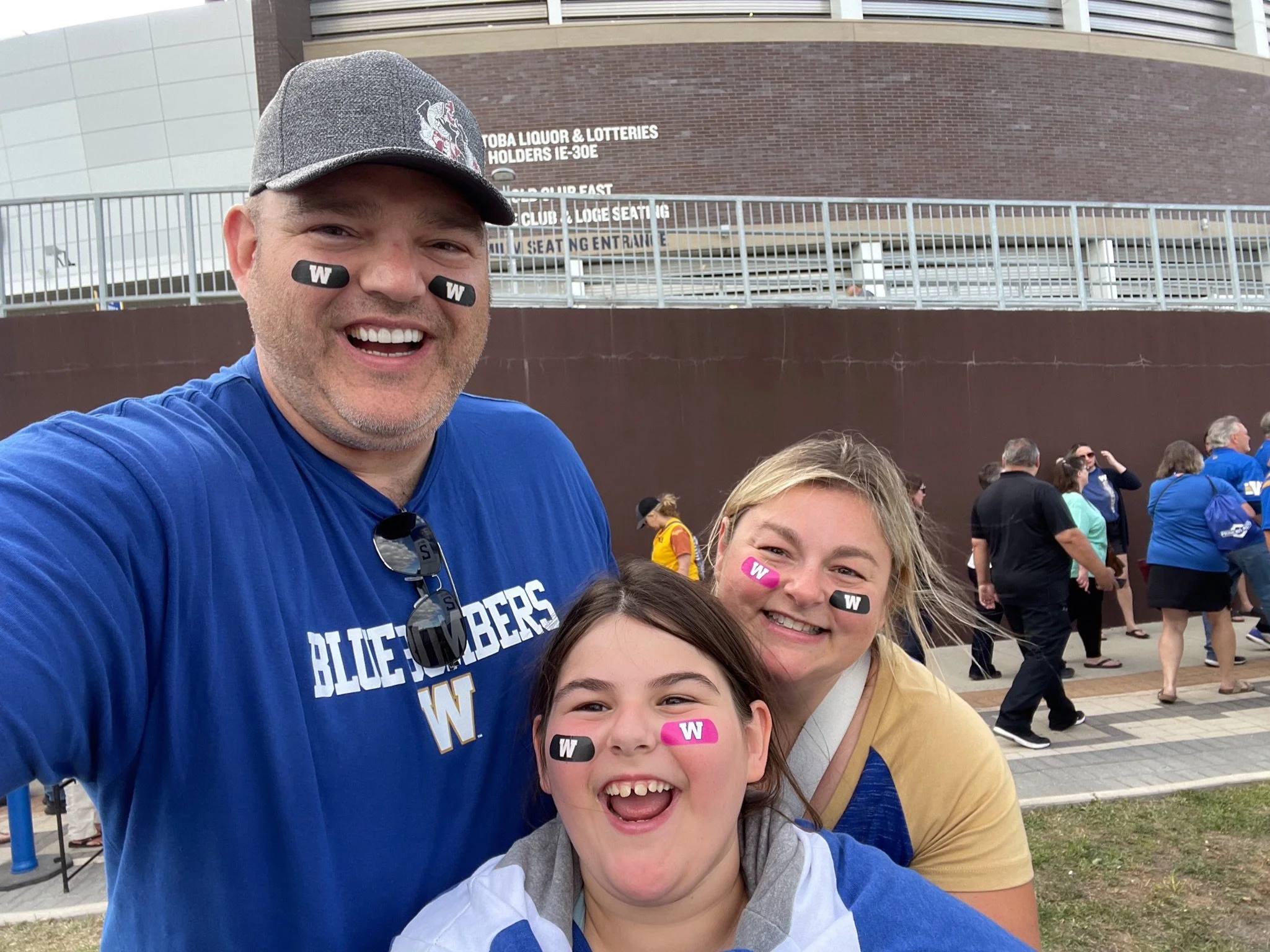 Laura with husband and daughter taking a selfie outside a large building with a crowd. The man wears a gray cap and blue sports shirt. At a football game.