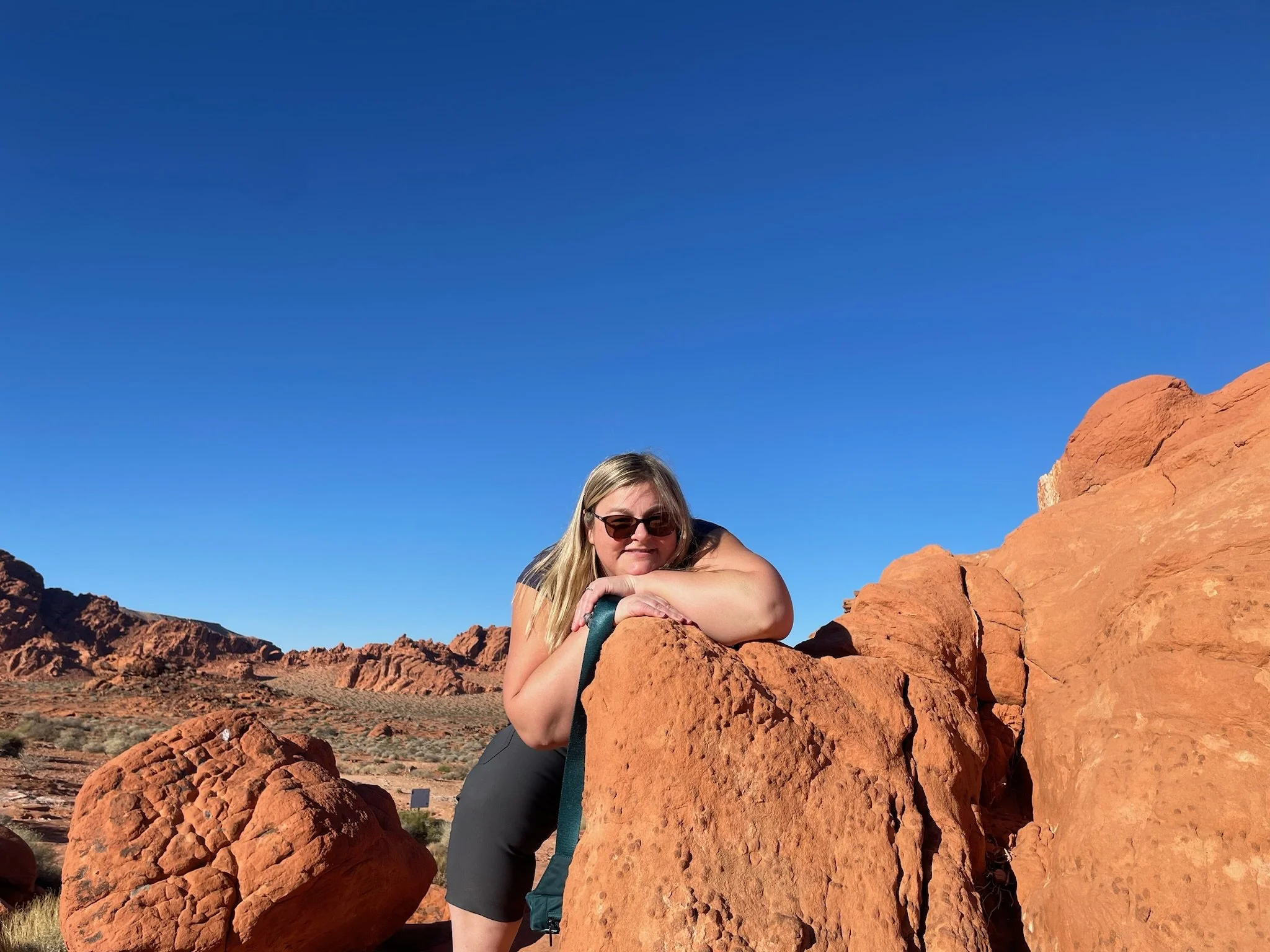 Laura, wearing sunglasses and black shirt, leaning on a large reddish rock formation in a desert landscape under a clear blue sky.