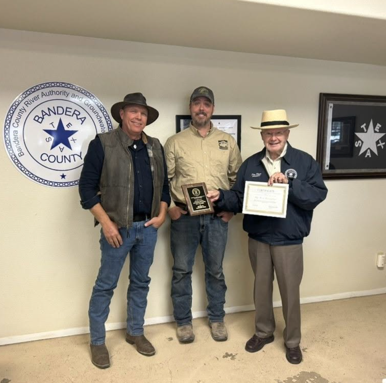 Three men standing inside a room, one of whom is holding a plaque and a certificate, in front of a wall with the Bandera County seal and a framed picture. All three men are smiling, winter-casual clothing, wearing hats.