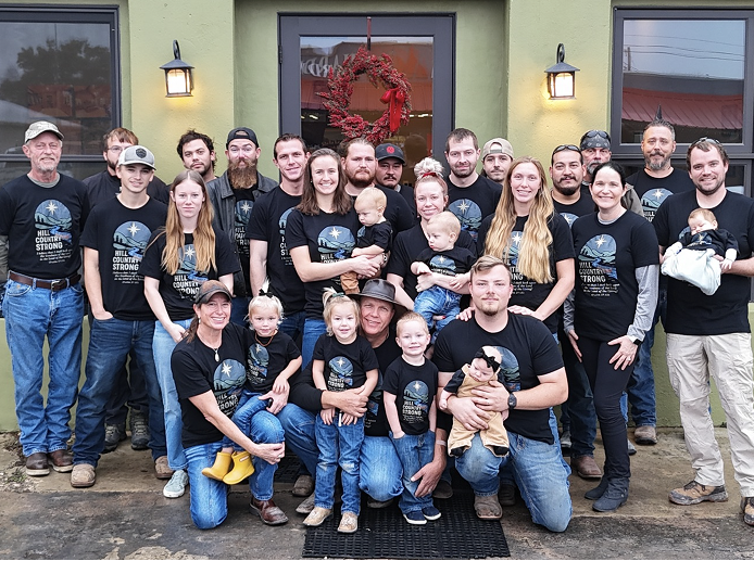 A large family gathering outside a house decorated for Christmas with a wreath and a red bow on the door. The group includes children, teenagers, adults, and seniors, most wearing matching black shirts with a moon and star logo that reads 'Hill Country Strong'. They are smiling and standing in front of a green exterior wall with black windows and exterior lights.