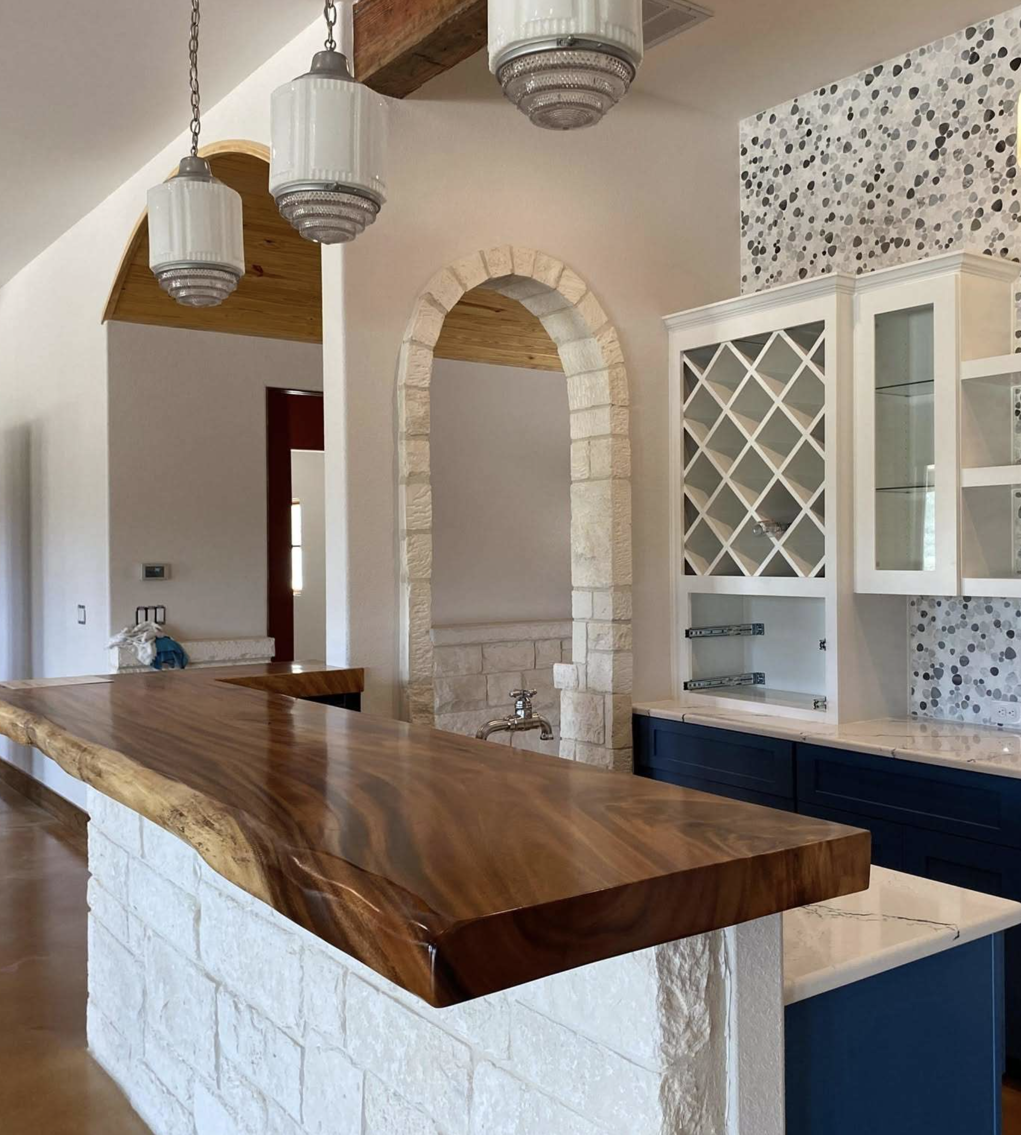 Interior view of a kitchen with a wooden countertop, white cabinets with glass doors, a stone archway, and three hanging pendant lights.