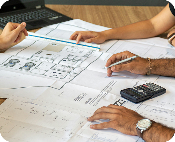 Two people working on architectural blueprints at a table, using a ruler and pen, with a calculator and a laptop nearby.