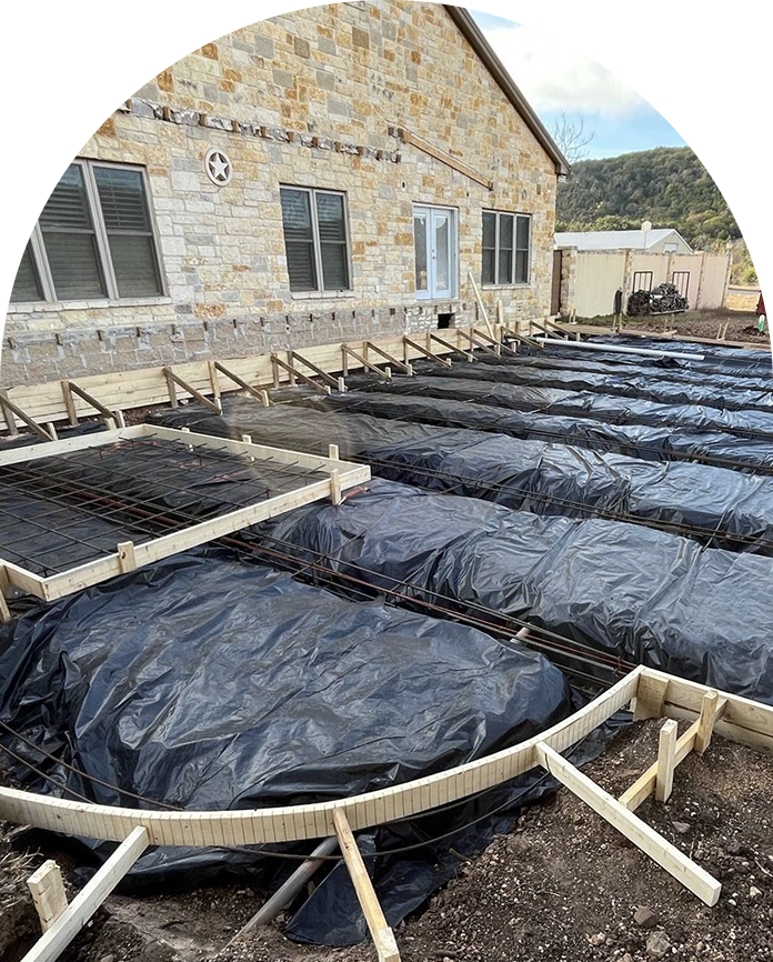 Construction site with foundation of a building, covered with black plastic sheets and surrounded by wooden frames, located next to a brick house with multiple windows.