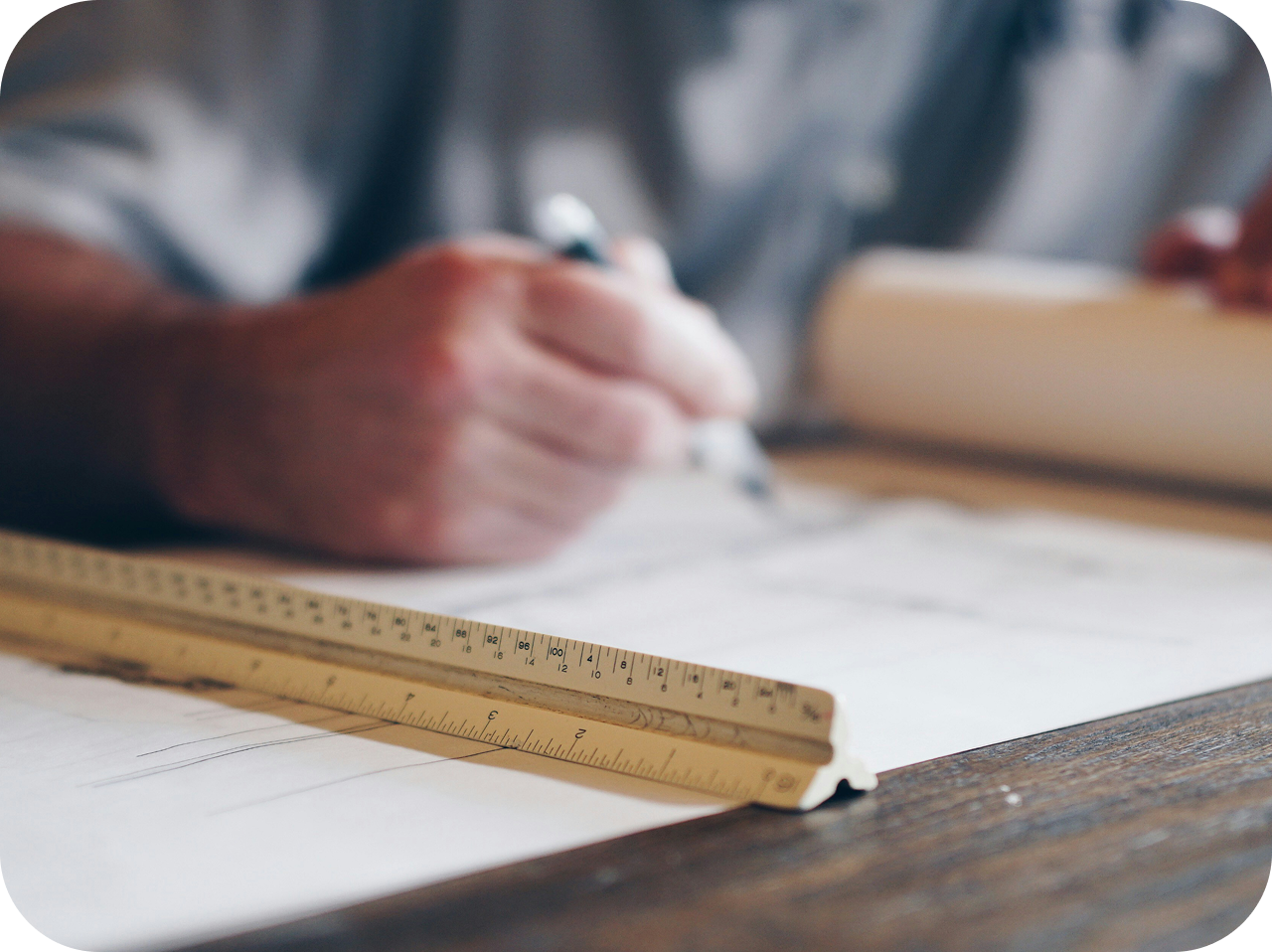 A person is writing on paper with a pen, with a ruler placed on the paper, on a wooden surface.