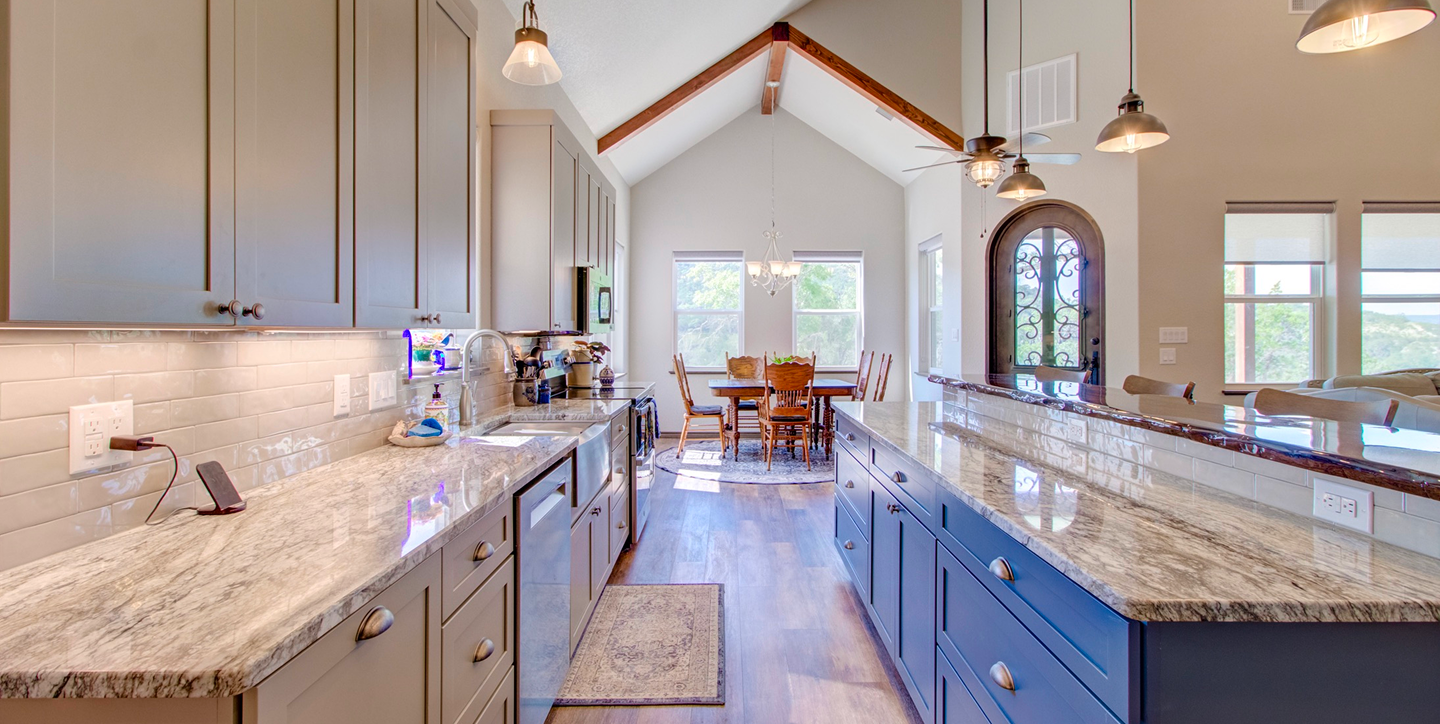 Kitchen with granite countertops, gray and blue cabinets, white backsplash, and hardwood floors. Open to dining area with a wooden table and chairs, large windows, and a ceiling with exposed beams and multiple hanging lights.