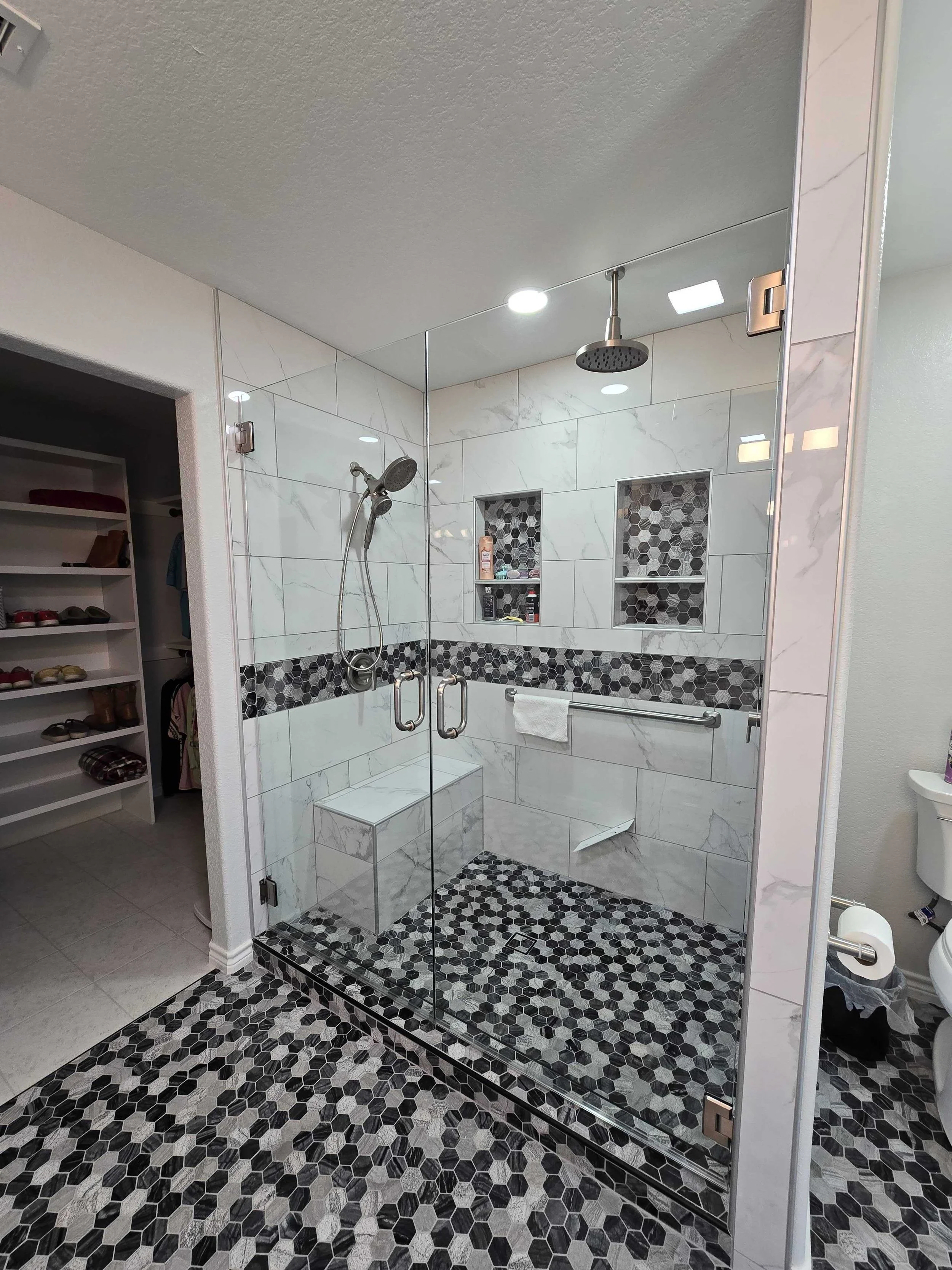 Shower with a glass door, black and white hexagonal tile floor, white marble wall tiles with black hexagon accents, two niches with toiletries, two showerheads, and a towel bar with a white towel.