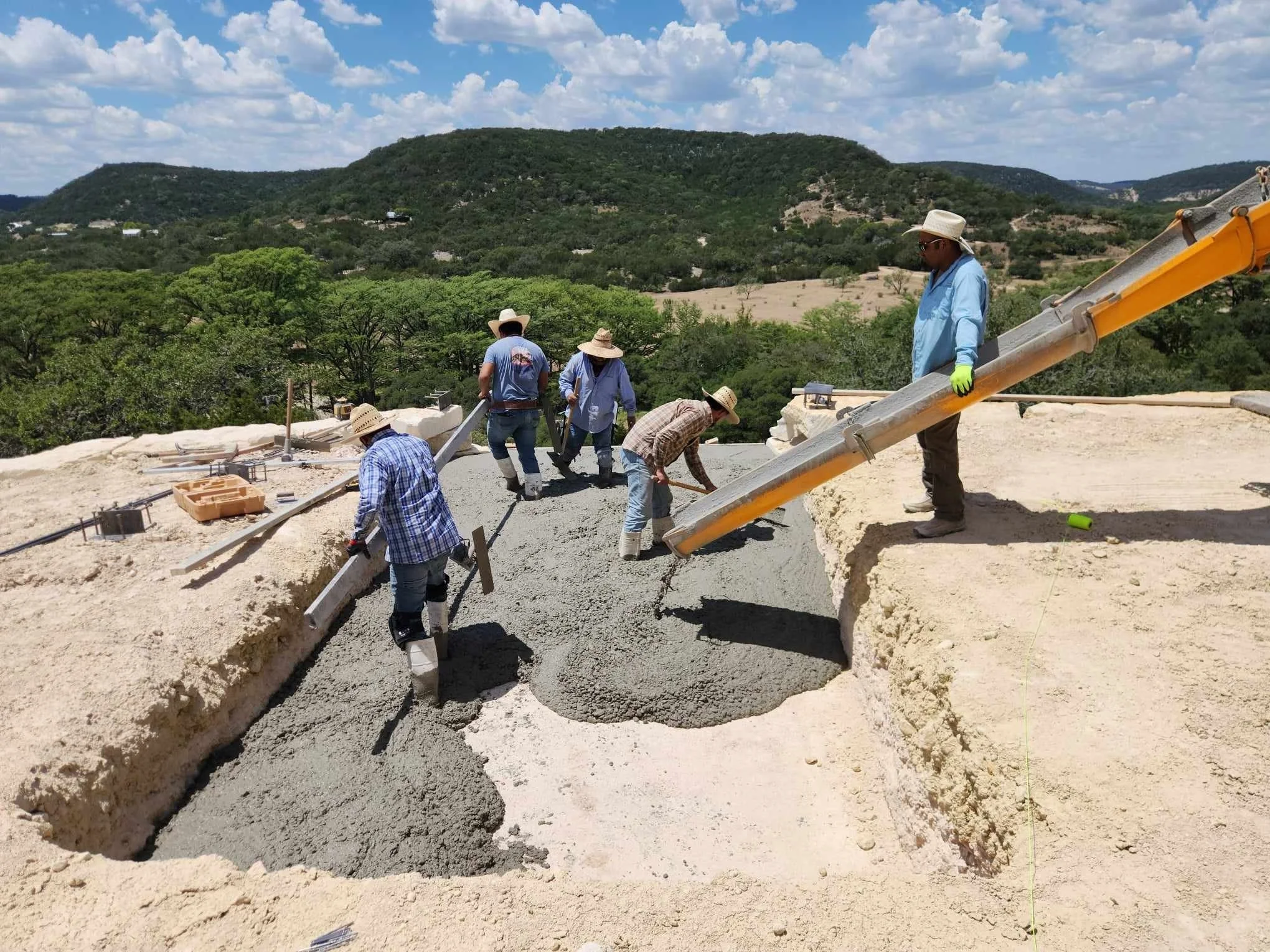 Construction workers pouring concrete for a building foundation, with a scenic landscape of hills and trees in the background.