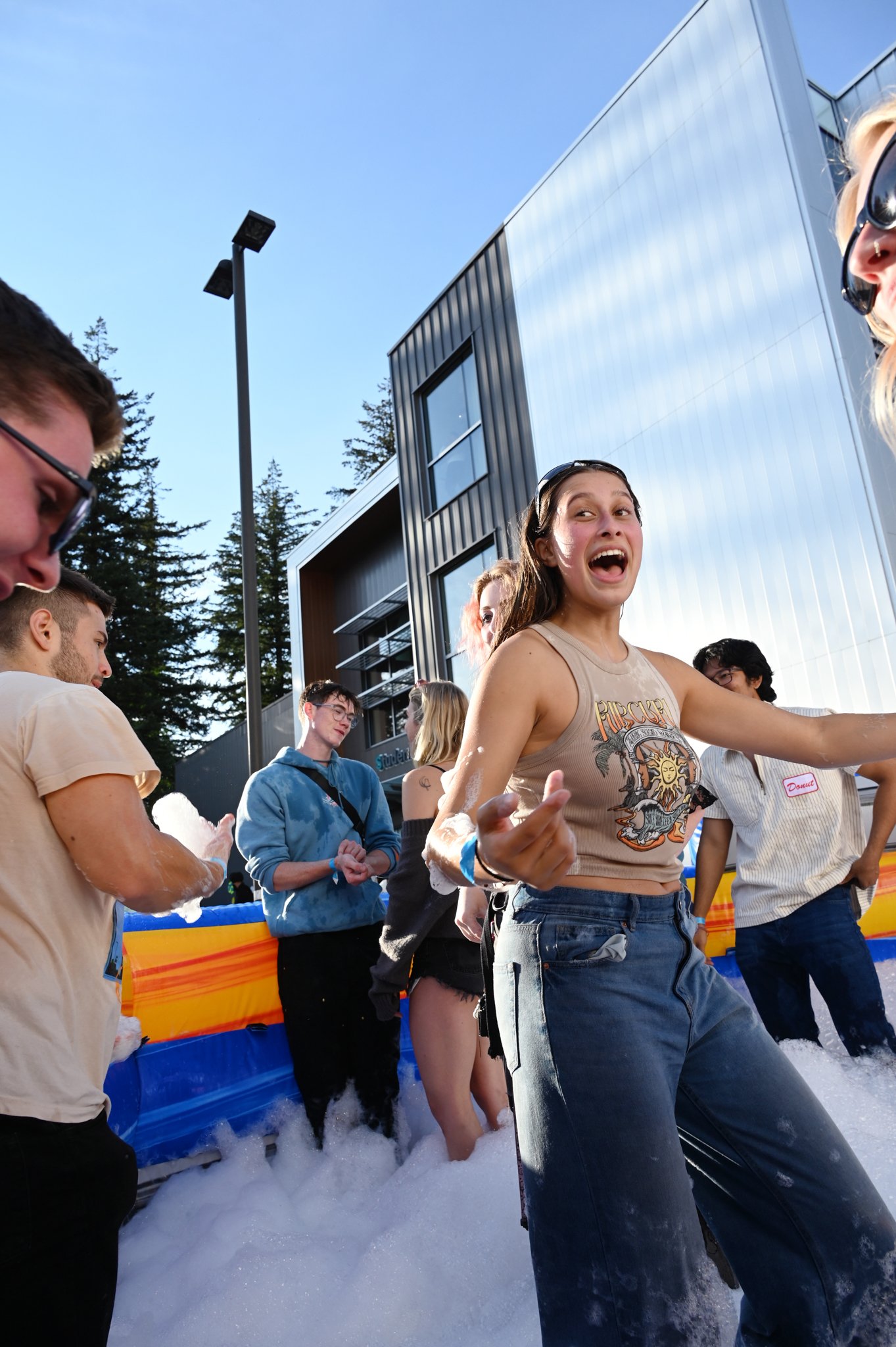 Group of young people having fun at an outdoor foam party, with foam on the ground and colorful inflatable barriers, in front of modern buildings and blue sky.