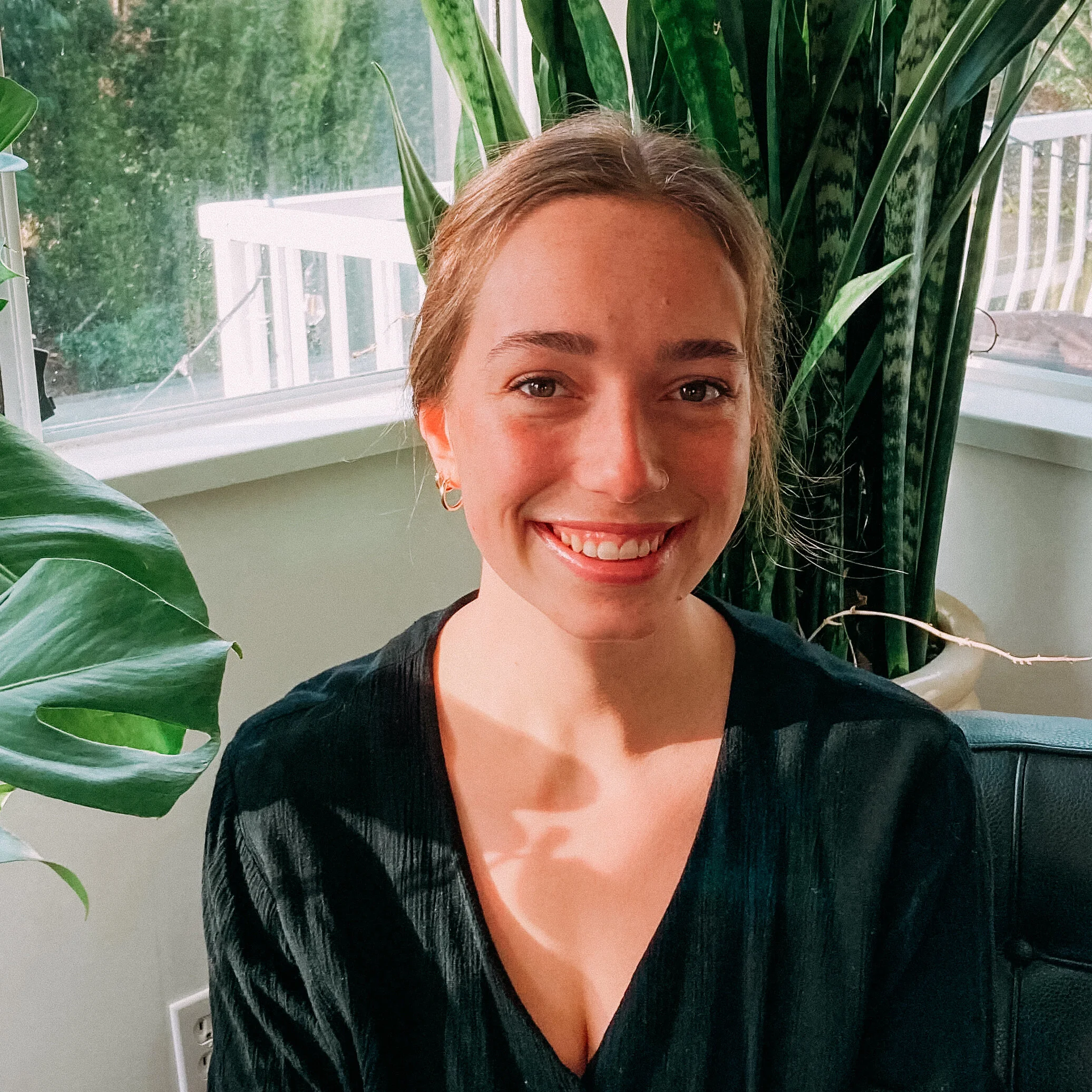 A woman is smiling in front of large green leafy plants and a window with a patio outside.