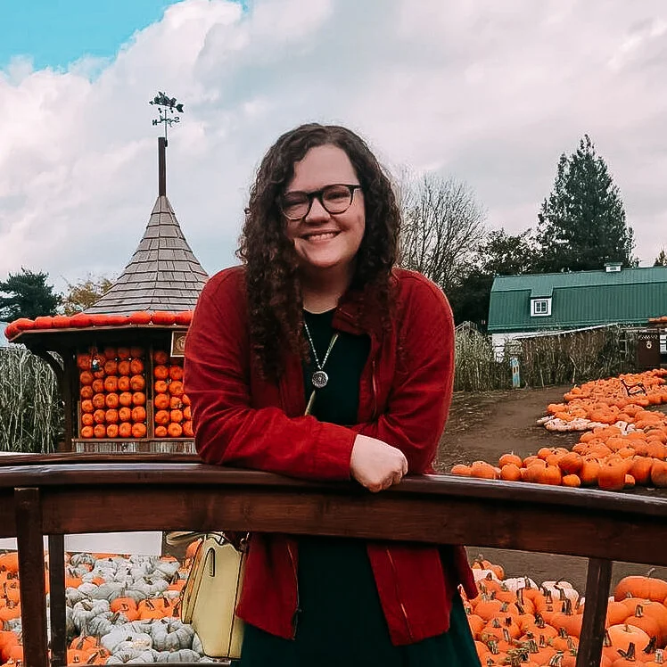 Woman with curly hair and glasses smiling, wearing a red jacket, leaning on a wooden fence surrounded by pumpkins at a pumpkin patch.