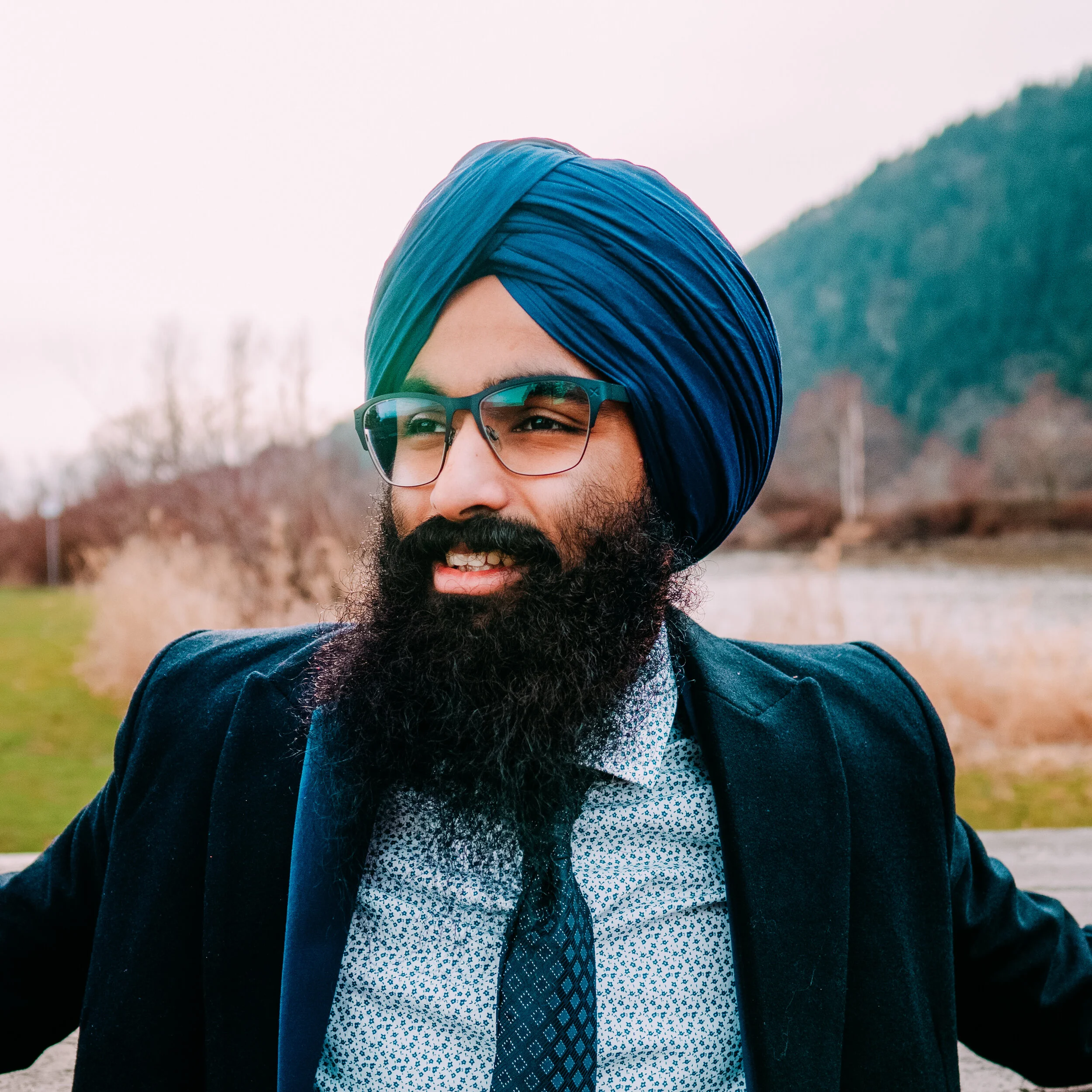 A person with a dark beard wearing glasses, a blue turban, a patterned shirt, a tie, and a suit jacket outdoors with trees and a mountain in the background.