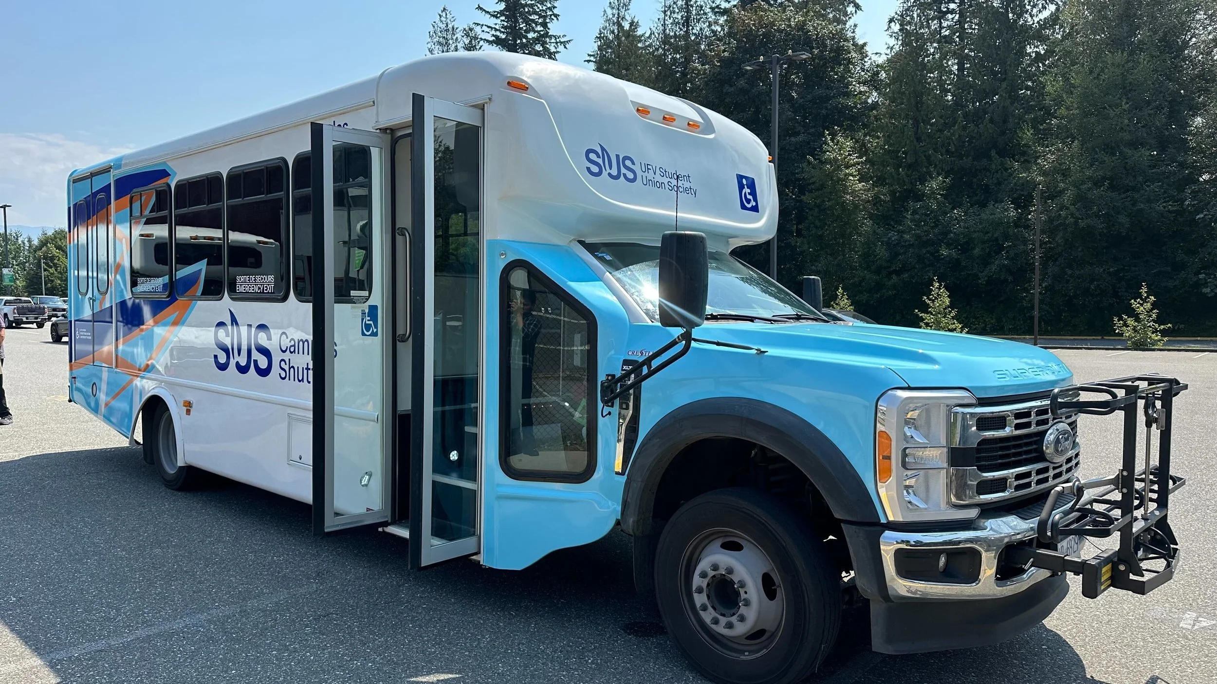 A blue and white bus with the text 'SUS Campus Shuttle' and 'UFV Student Union Society' on its side, featuring accessibility symbols, parked in an outdoor lot with trees in the background.