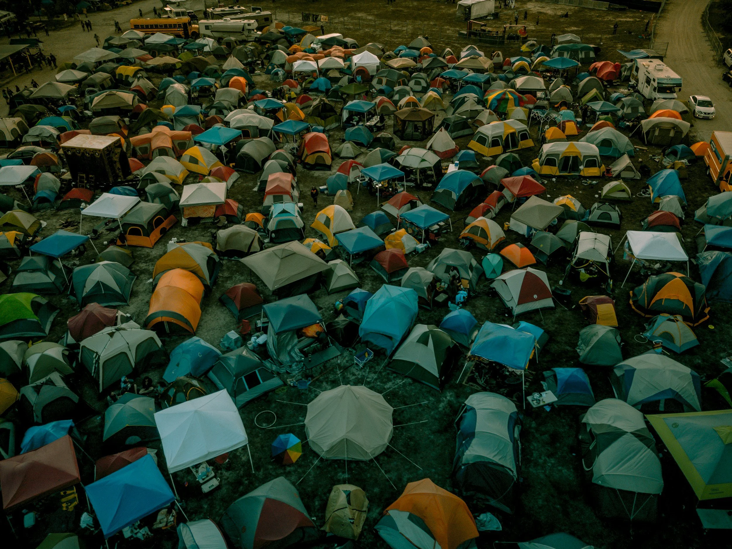 Aerial view of a large campsite with numerous tents in various colors, surrounded by parked vehicles and trailers. A dirt road curves around the campsite, with some people visible walking or standing.