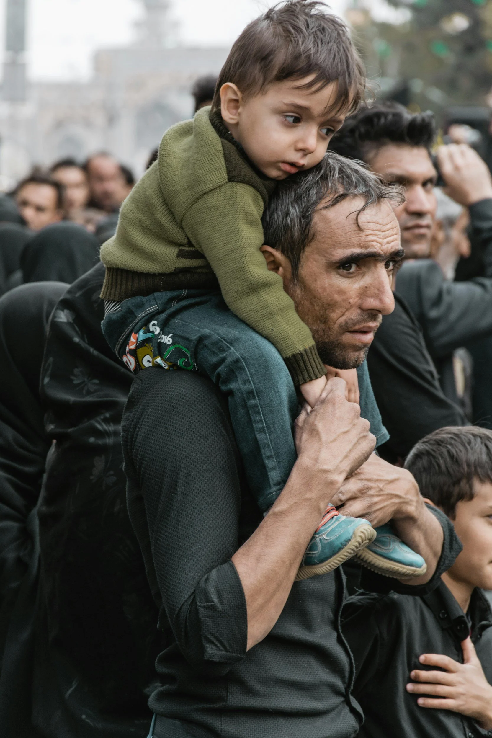 A man with gray hair and a beard is holding a young boy on his shoulders during a crowded outdoor event. The boy looks to the side with a serious expression, while the man appears focused and protective. Several other people are visible in the background.