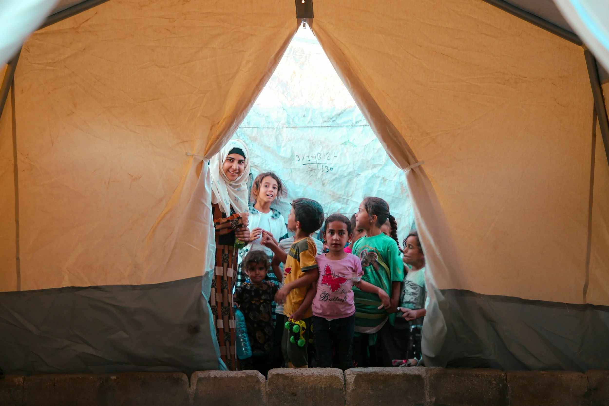 People, mostly children, gather inside a tent, with some smiling and looking toward the camera, while the photo is taken from outside the tent through its opening.