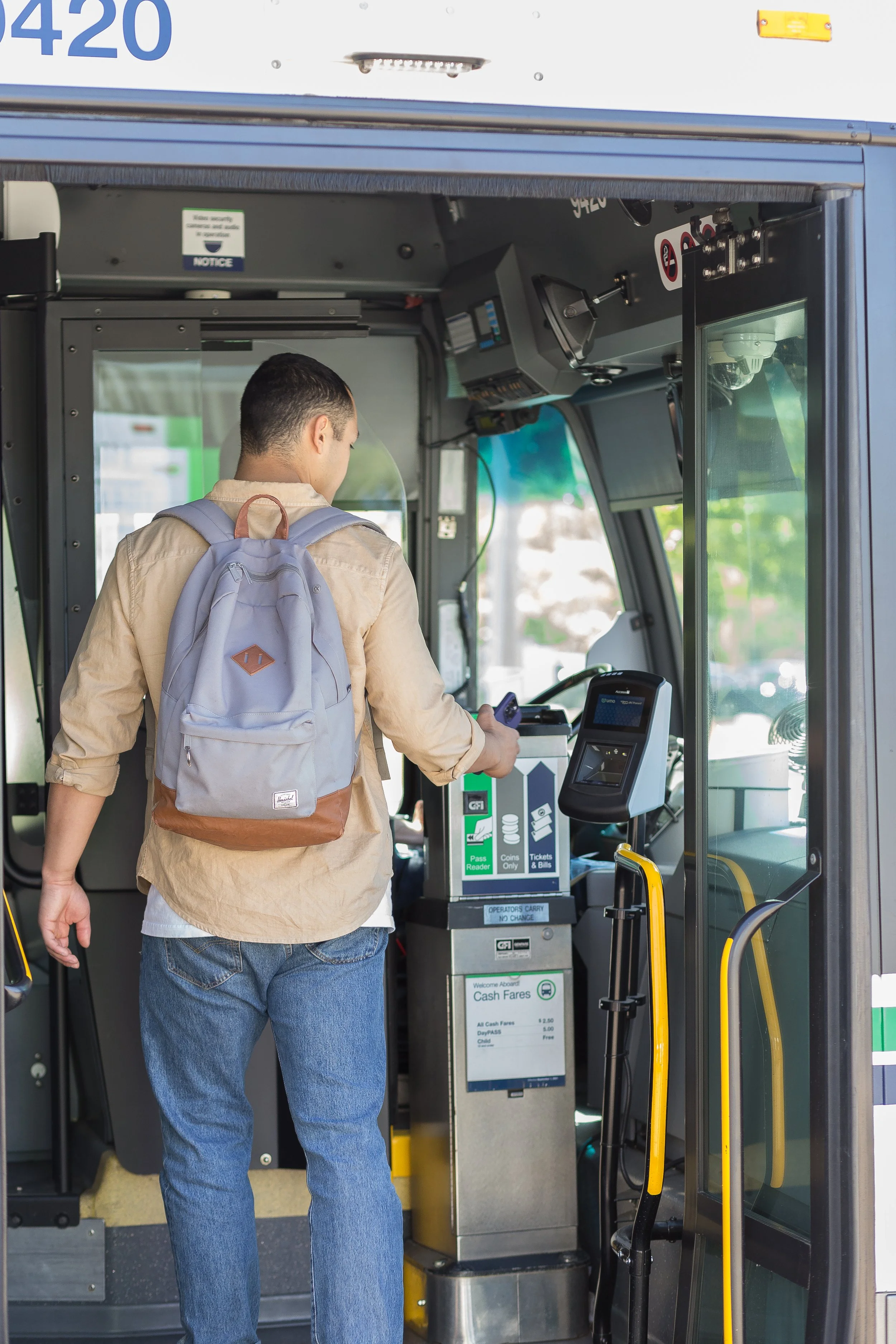 A man with dark hair carrying a light purple backpack, wearing a beige shirt and blue jeans, using a contactless payment terminal at a bus fare machine inside a bus, with controls and signage visible.