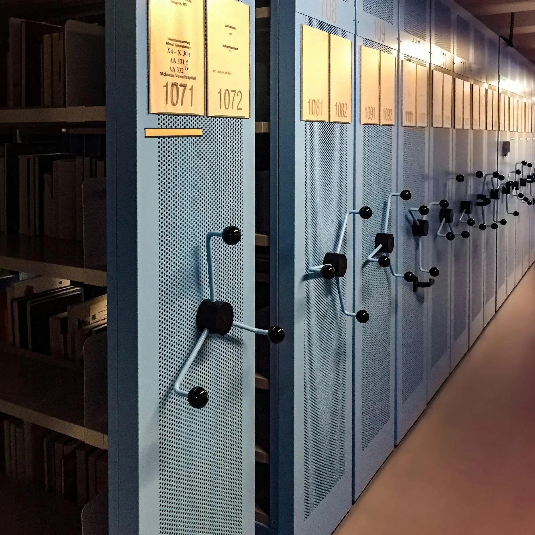 A row of gray and blue lockers with yellow labels displaying numbers, located in a library or similar setting, with bookshelves partially visible on the left