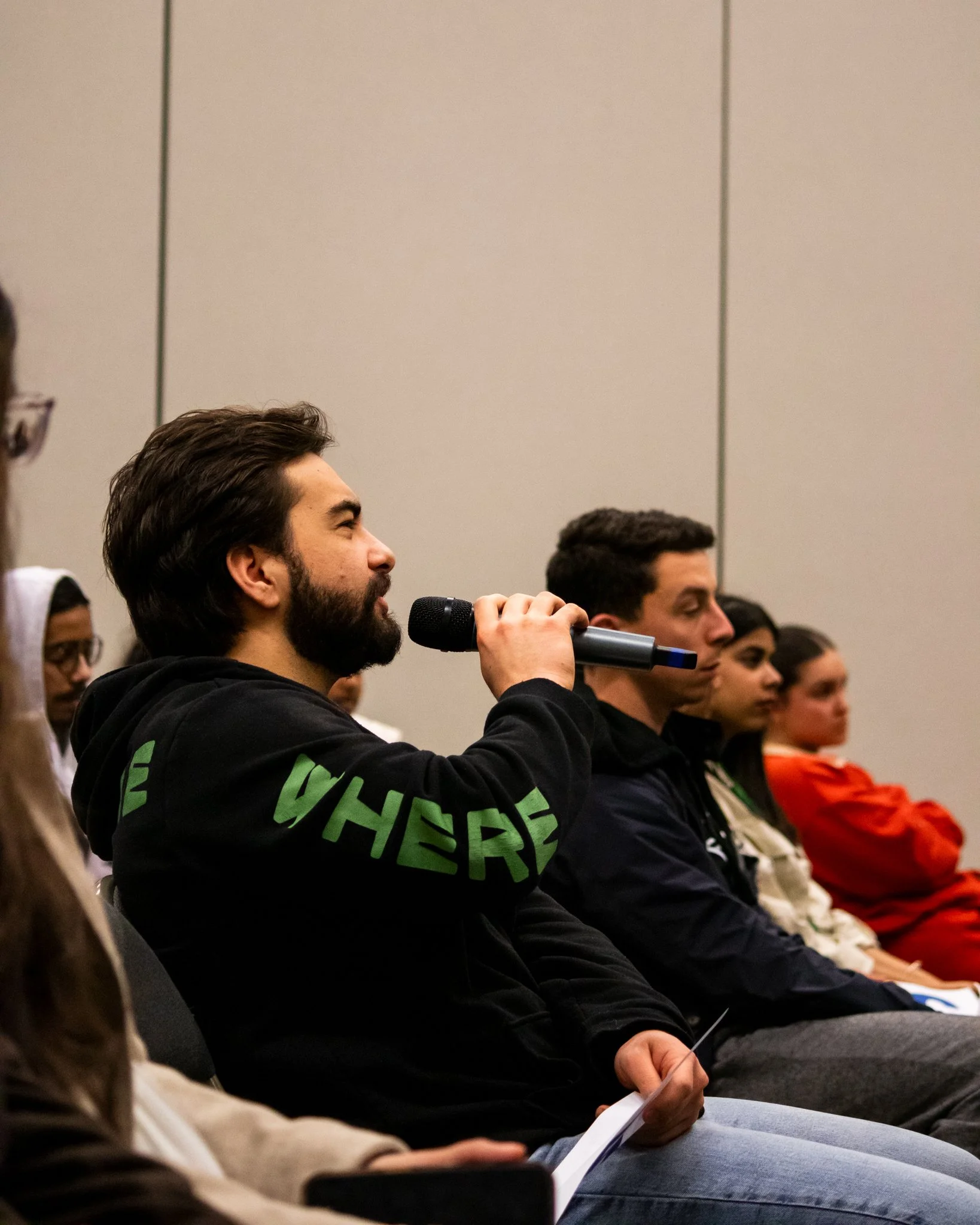 A man with brown hair and a beard, wearing a black hoodie with green text on the sleeve, is holding a microphone and speaking in a seated audience. Several other people are sitting next to him, listening attentively in a room with gray walls.