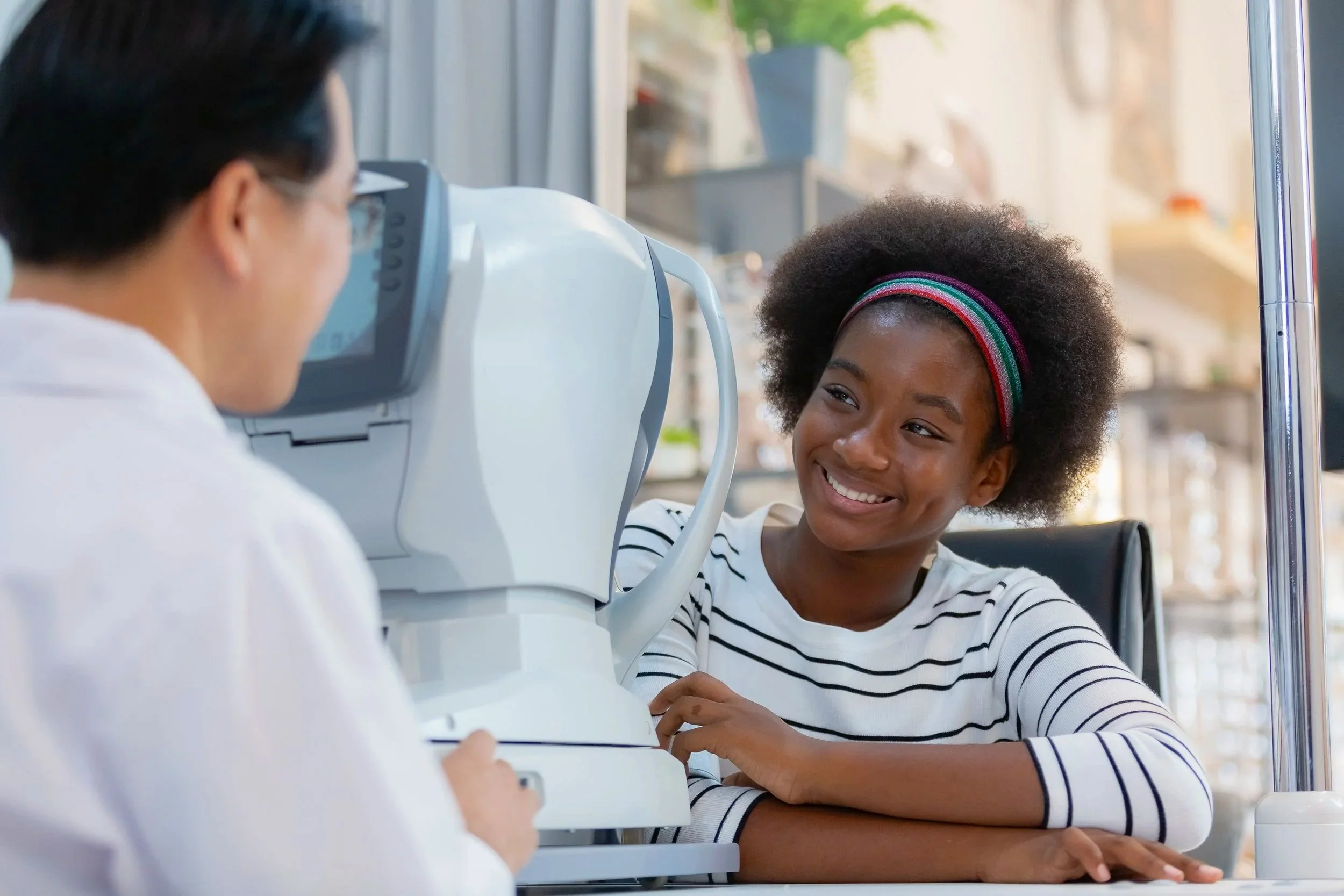 A young Black girl in a striped shirt smiling at an eye doctor during an eye examination, with medical equipment and a neutral background visible.