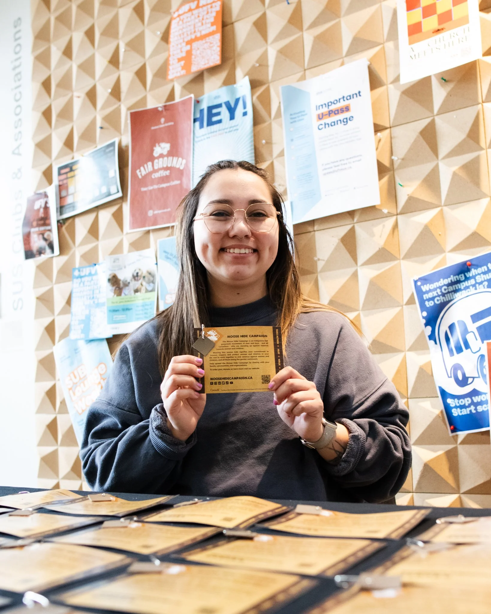 A young woman with glasses smiling while holding a yellow campaign flyer at an indoor event. Behind her, a bulletin board displays various posters and flyers related to social causes and campaigns.