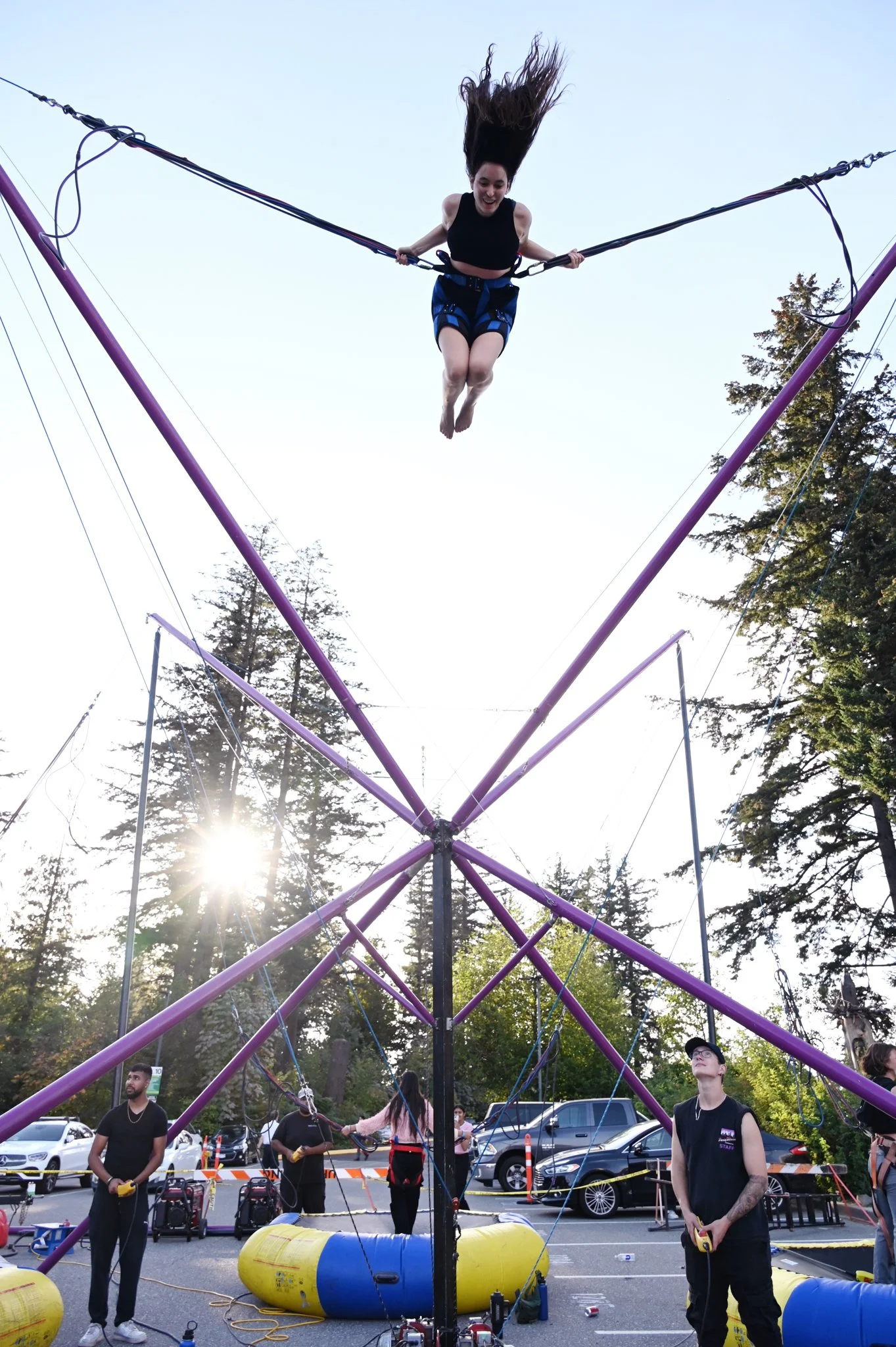 A woman with long hair and a black crop top is bouncing on a large trampoline outdoors, with her hair flying upwards. Several people are standing around the trampoline, holding controllers, and a safety barrier surrounds the area. There are trees and parked cars in the background.