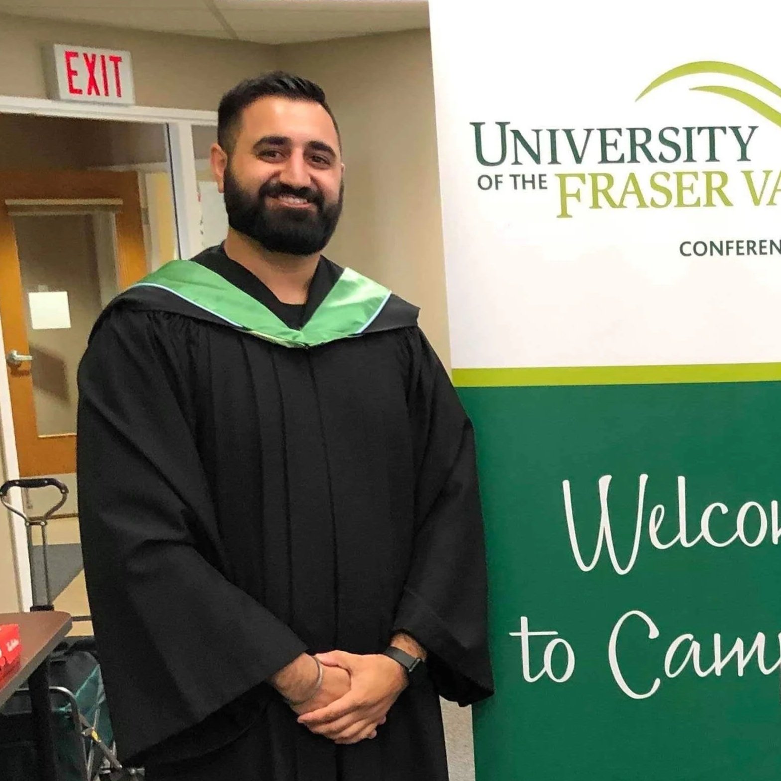 A man in a graduation cap and gown standing in front of a sign for the University of the Fraser Valley, smiling with arms crossed.