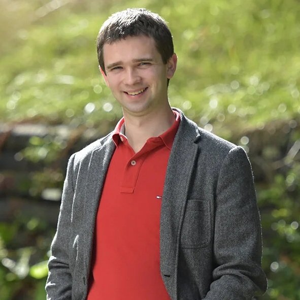 A smiling man with short dark hair in a red polo shirt and gray blazer, standing outdoors with a blurred green background.