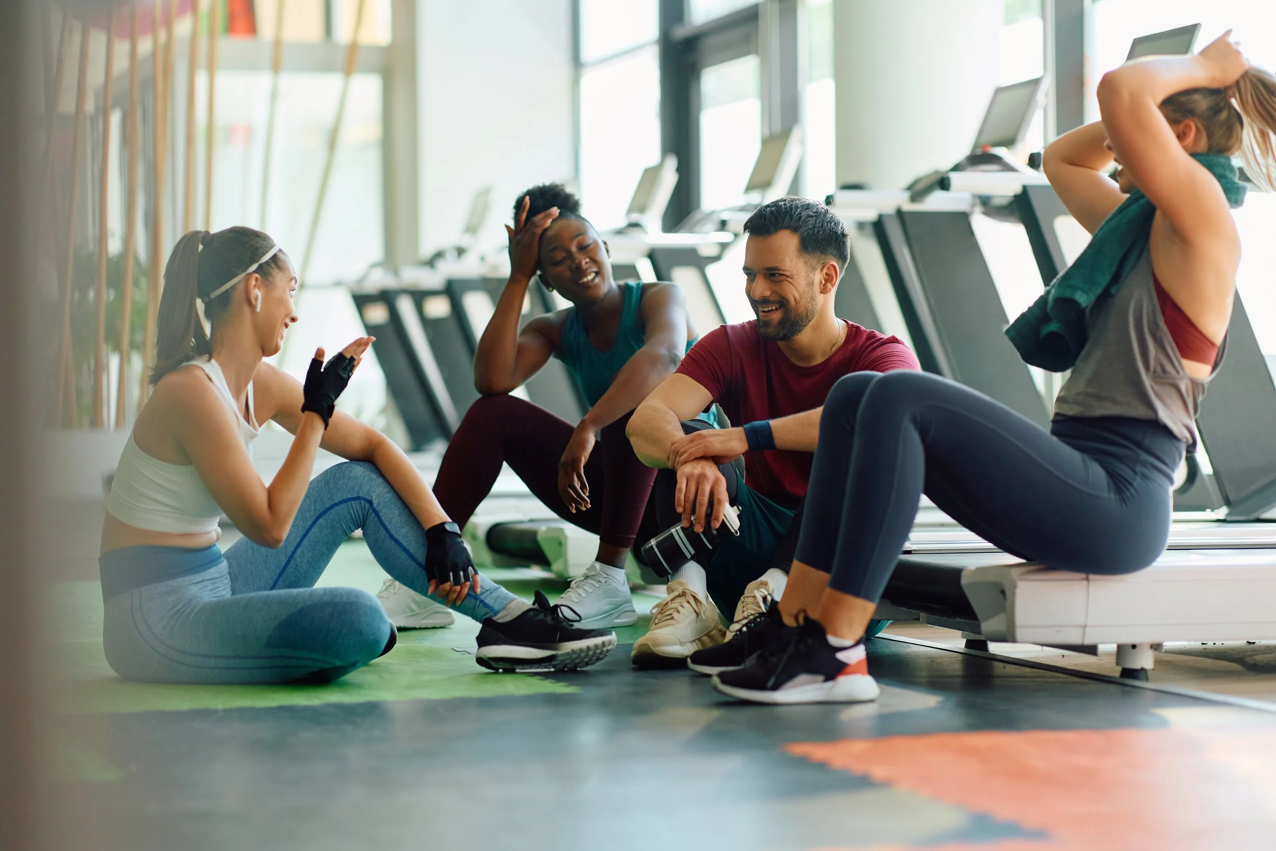 Four people resting after a workout at the gym, sitting on the floor near treadmills, laughing and chatting.