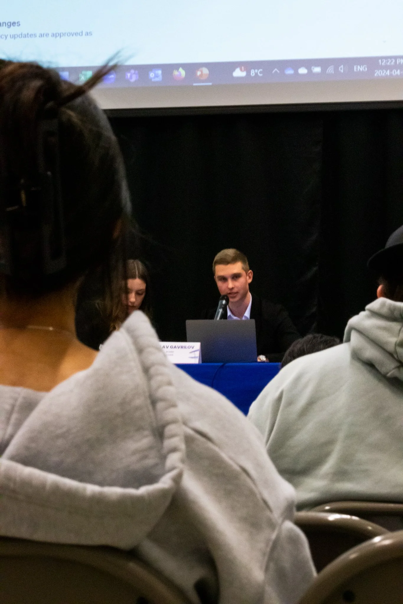 A person in a white hoodie sitting in an auditorium or conference room, facing forward, with many chairs and other people around. A young man is speaking or presenting at a table with a laptop.
