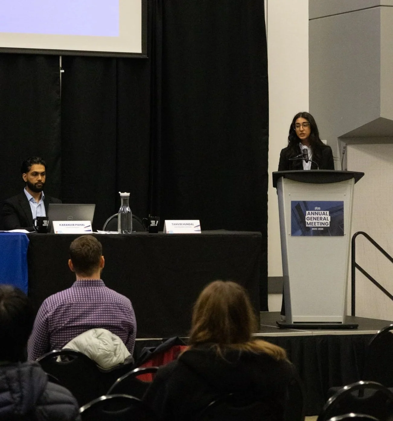 A woman is speaking at a podium during an annual general meeting, with two men seated at a table on stage and audience members in front.