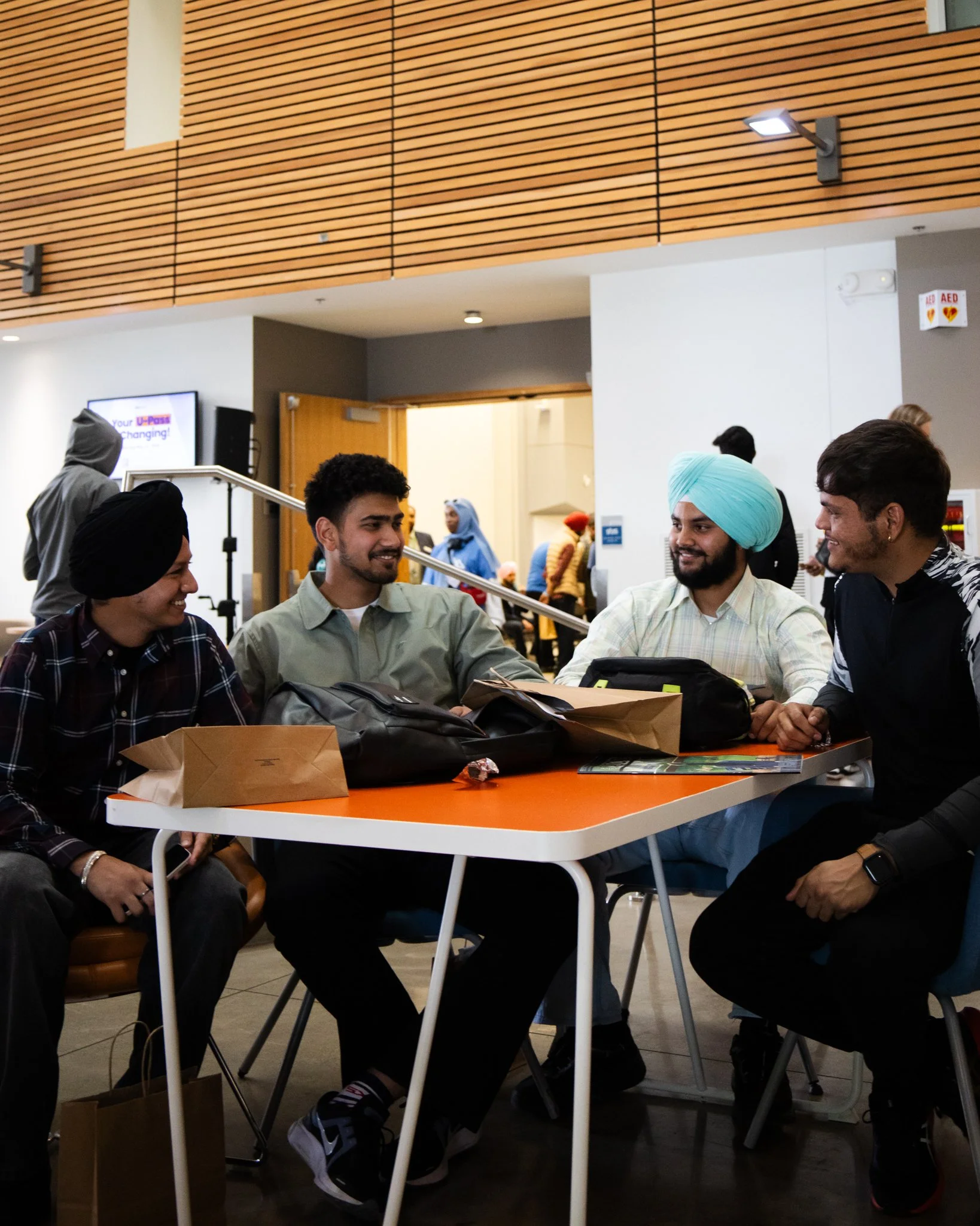 Group of young adults sitting at a table, engaged in conversation inside a modern building with wooden wall panels and overhead lighting.