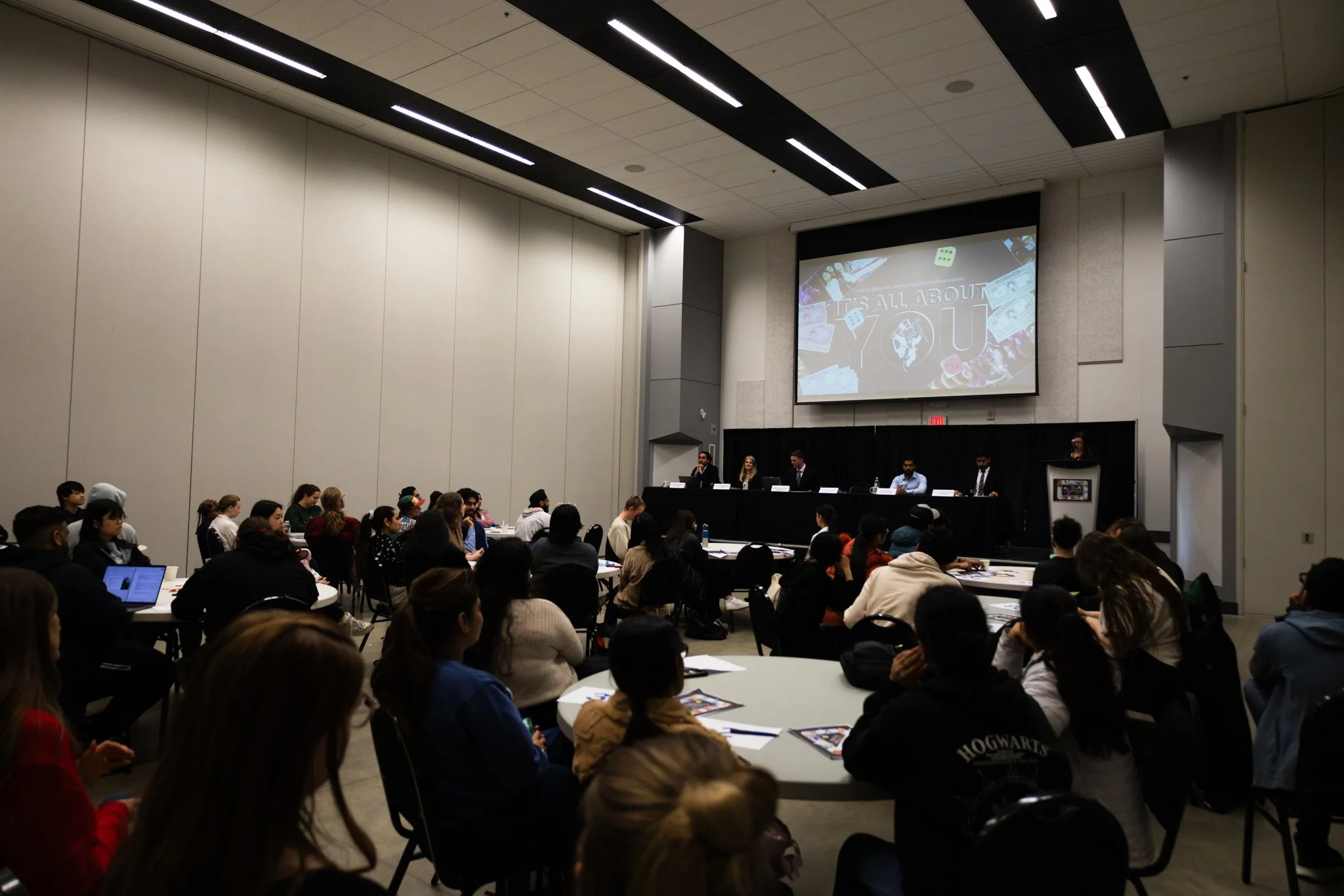A large conference room filled with people sitting at tables, listening to a panel of five individuals seated at a long table at the front. A large screen behind the panel displays a presentation titled 'IT'S ALL ABOUT YOU' with images of money, playing cards, and dice.