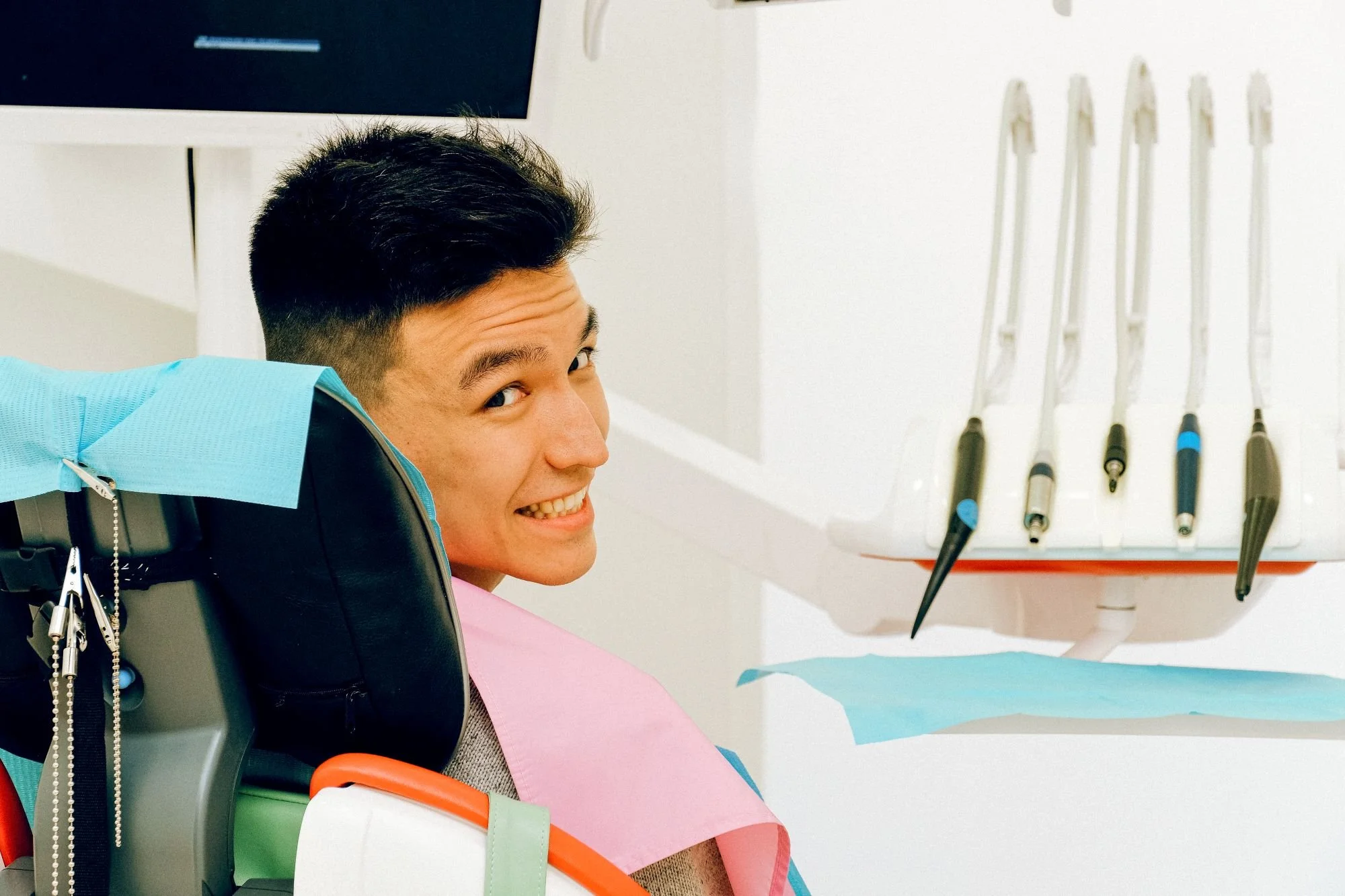 A young man smiling in a dental chair with colorful dental bibs, with dental tools hanging on the wall behind him.