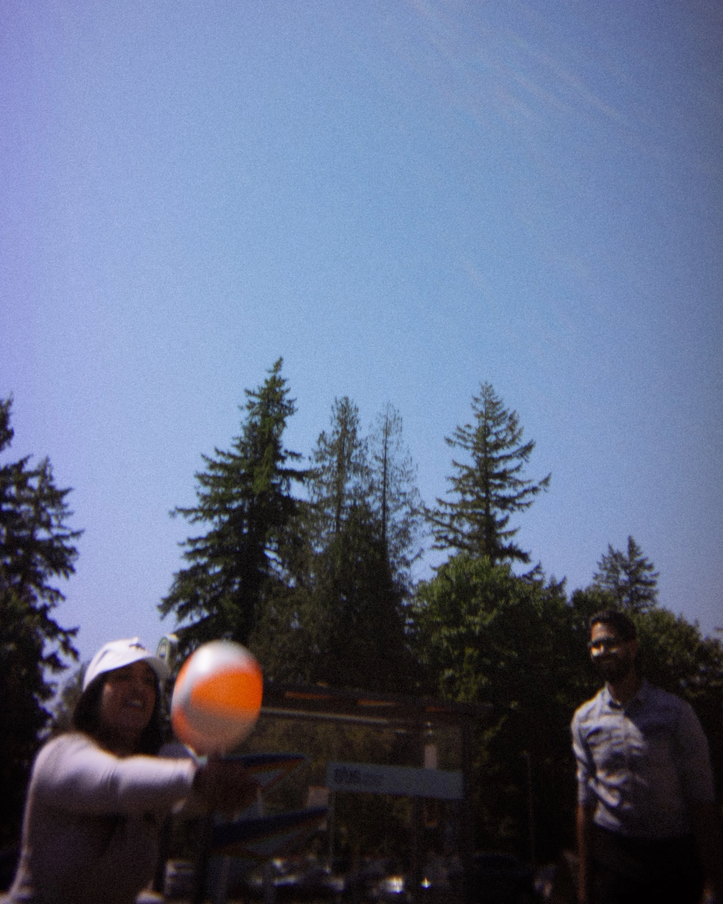 Group of children outdoors at dusk, some wearing helmets, facing a display table with a sign, surrounded by tall trees, with a clear sky overhead.