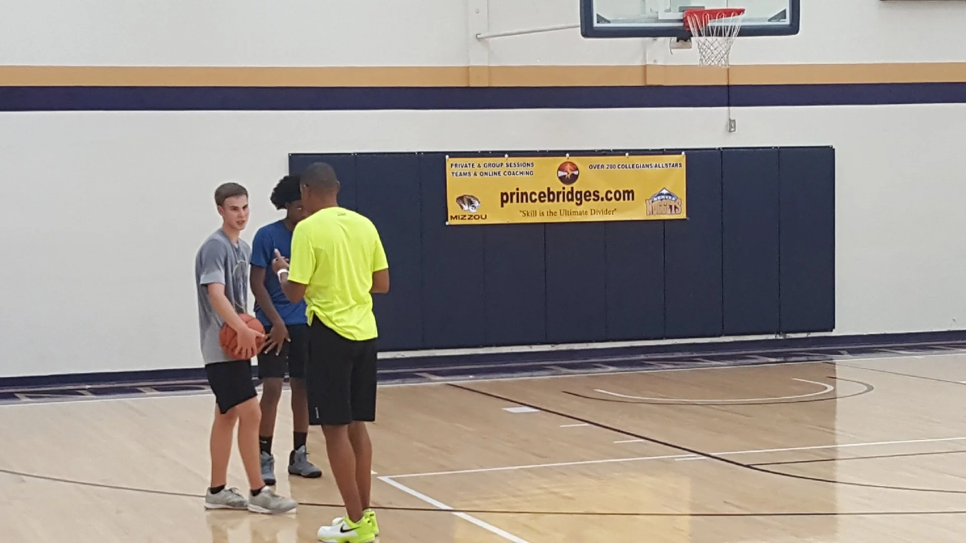 Three young male basketball players talking inside a gymnasium, with a yellow banner advertising princebridges.com in the background.
