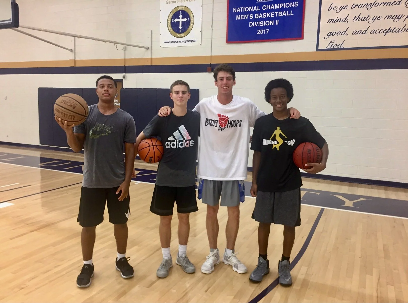 Four young men standing together on a basketball court, holding basketballs. They are smiling and posing for the photo. The gym has banners on the wall, including one for a basketball division championship in 2017.
