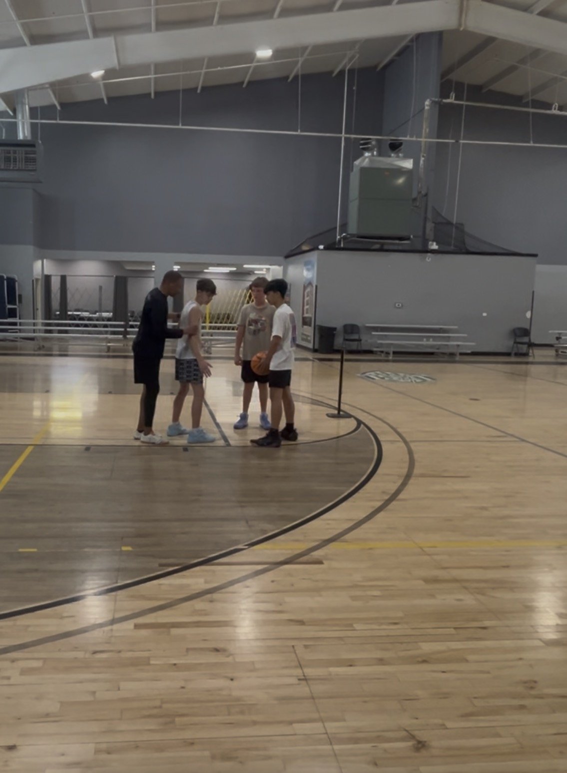 Four young men standing on a basketball court inside a large gym or sports arena, engaged in conversation.