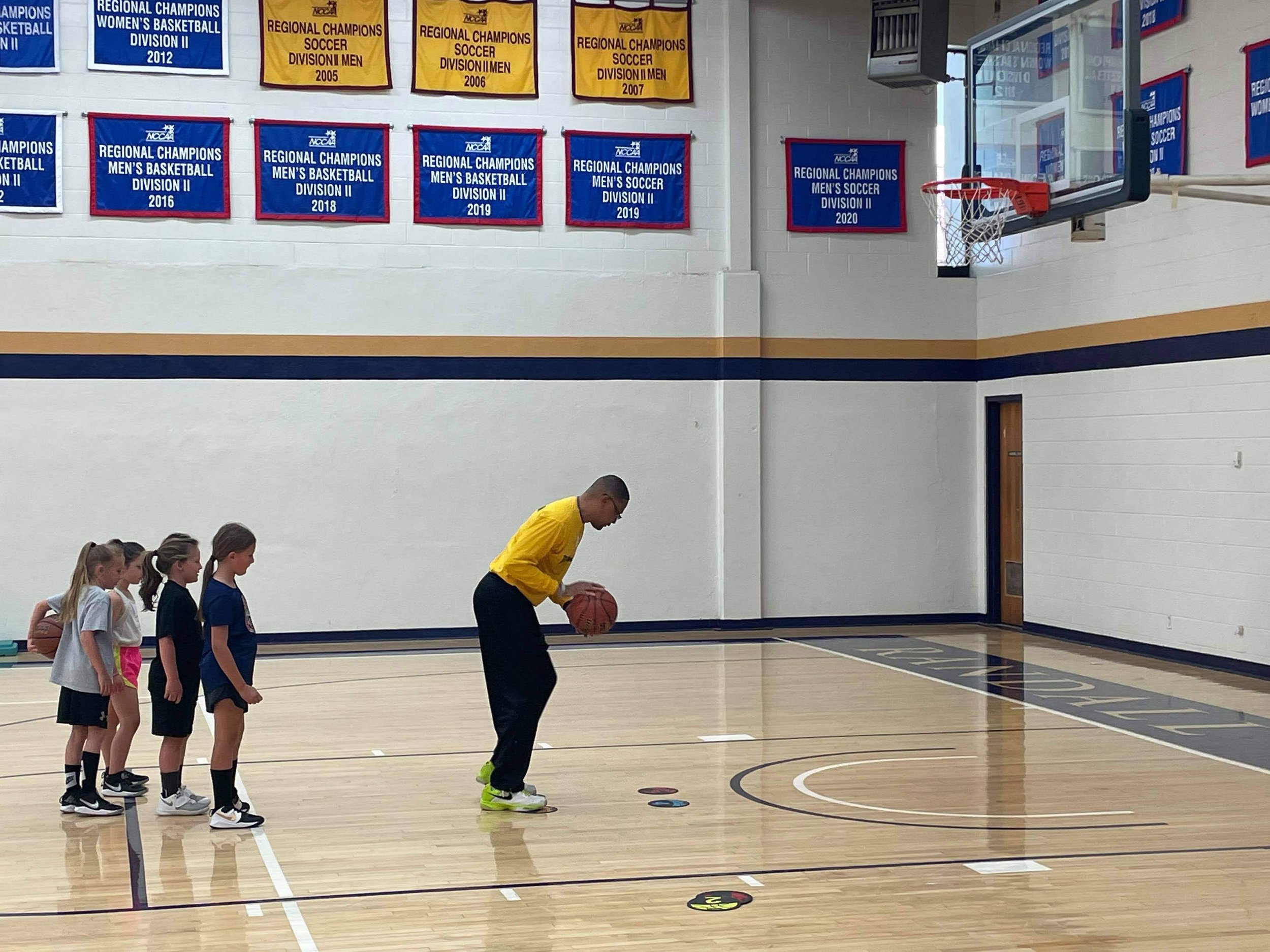 A group of young girls lined up in a gymnasium, each holding a basketball and preparing to dribble or practice. An adult coach in a yellow shirt is demonstrating a basketball drill. The gym wall is decorated with banners celebrating athletic achievements and championships.