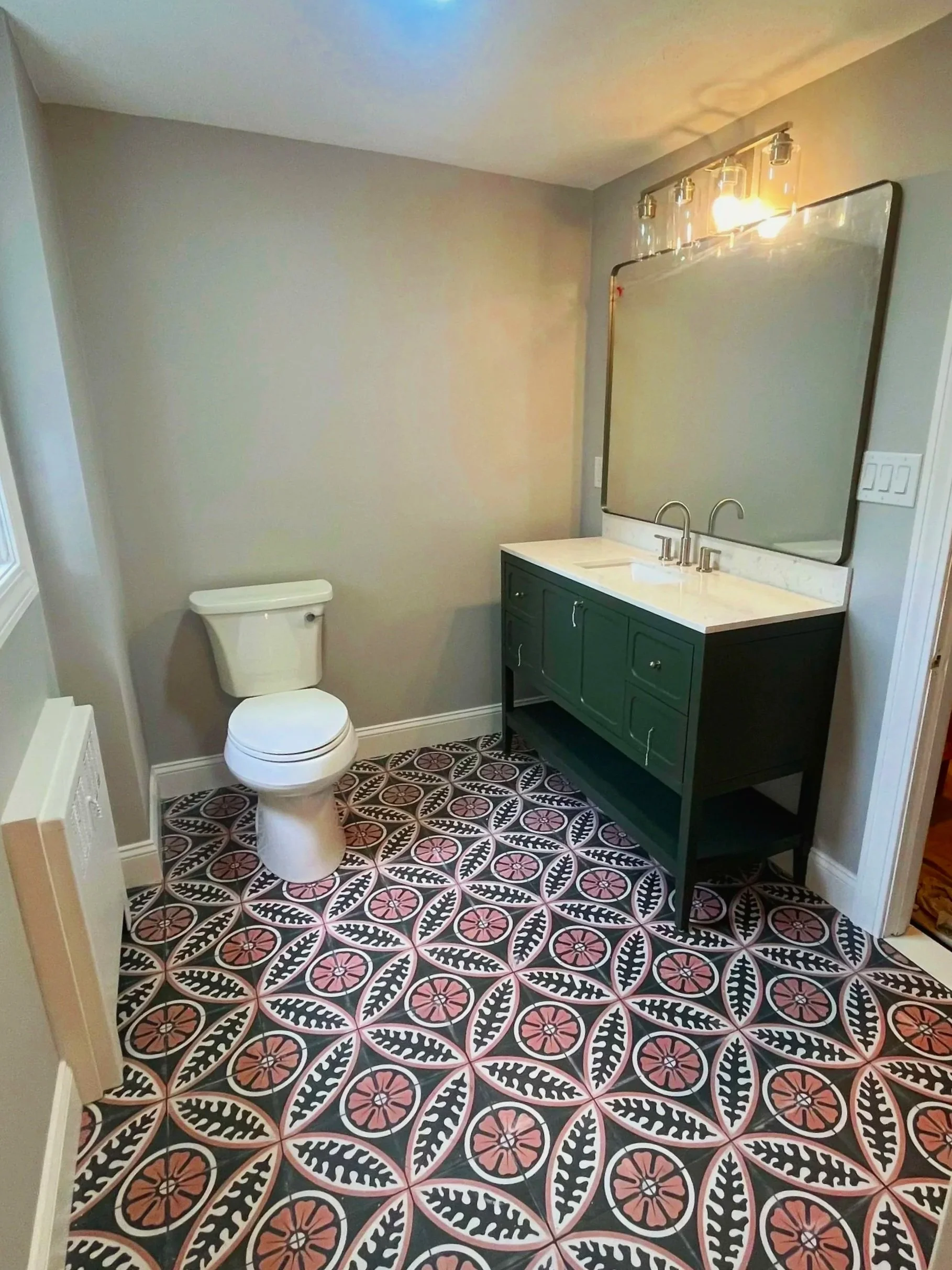 A small bathroom with a toilet and a green vanity with a marble countertop, a large rectangular mirror, and a patterned tile floor with floral designs in black, white, pink, and beige.