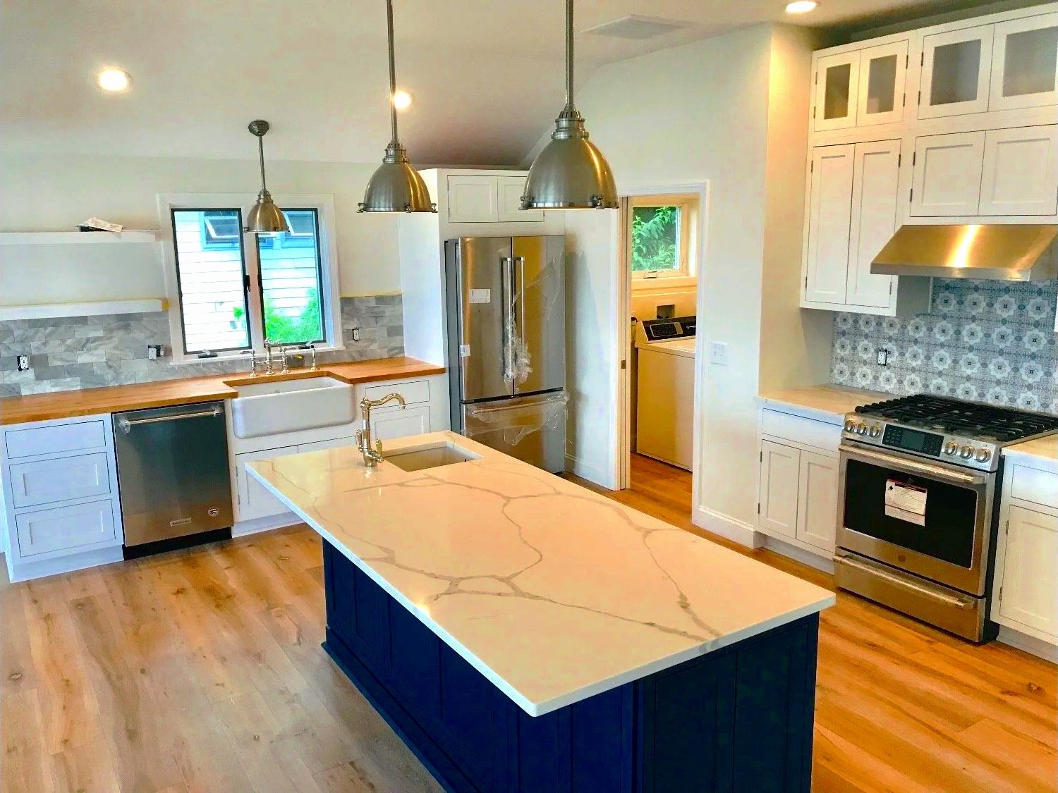 Modern kitchen with white cabinetry, a large kitchen island with a marble countertop, stainless steel appliances including a refrigerator, oven, dishwasher, and range hood, pendant lighting, a window above the sink, and hardwood floors.