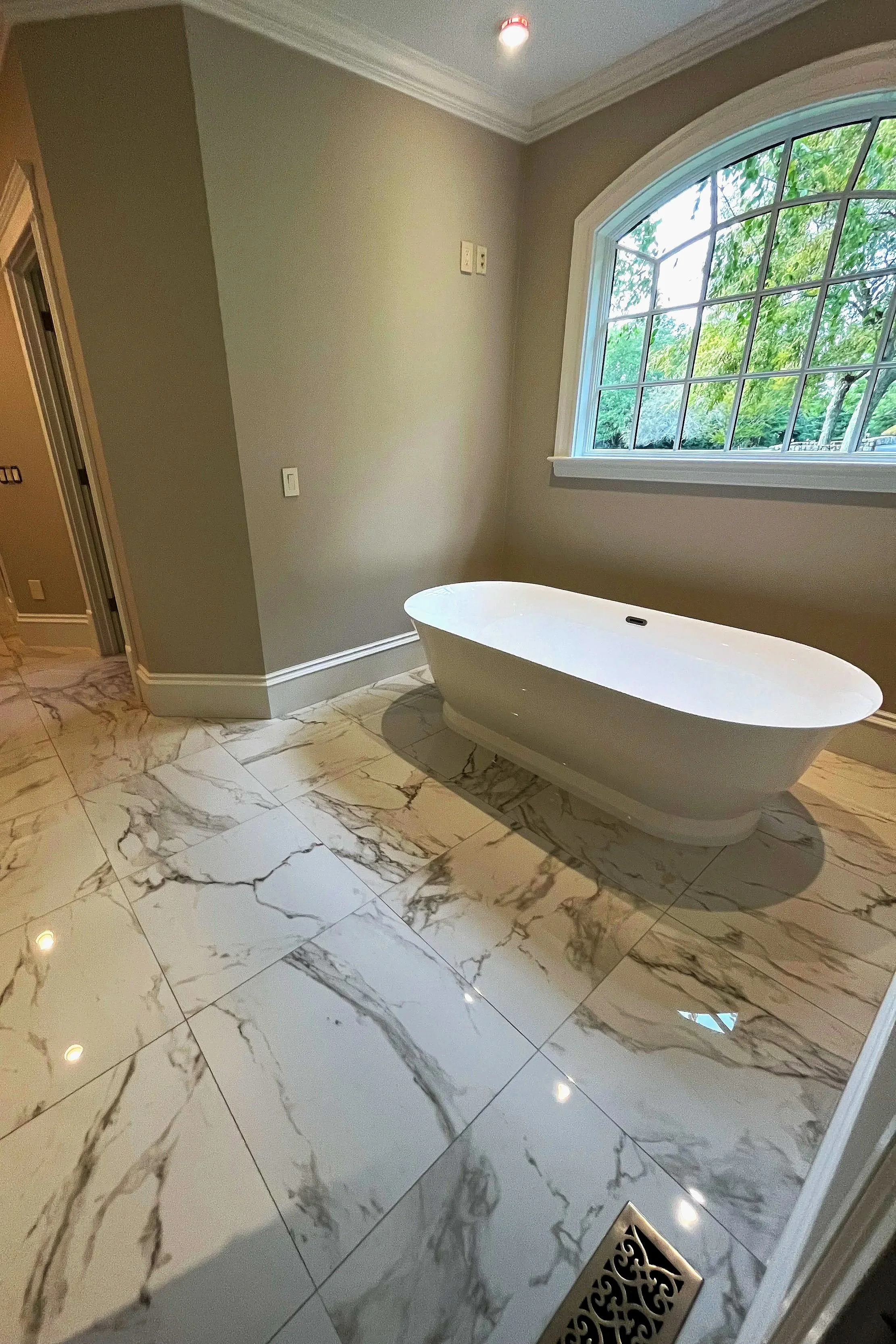 A bathroom with a white freestanding bathtub, marble tile flooring, a large arched window with green trees outside, and a decorative floor drain.