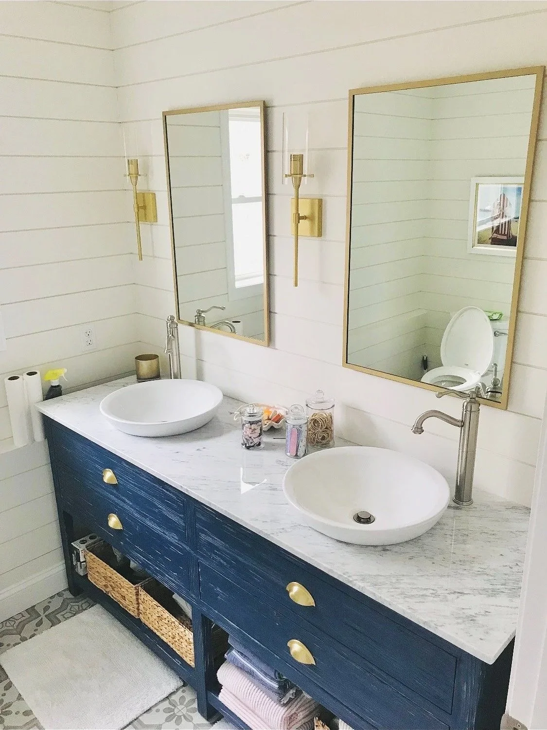 Bathroom with a double vanity featuring two vessel sinks, a marble countertop, and gold hardware. There are two mirrors, gold wall sconces, and decorative jars on the counter. The floor has patterned tiles, and there is a small white rug in front of 